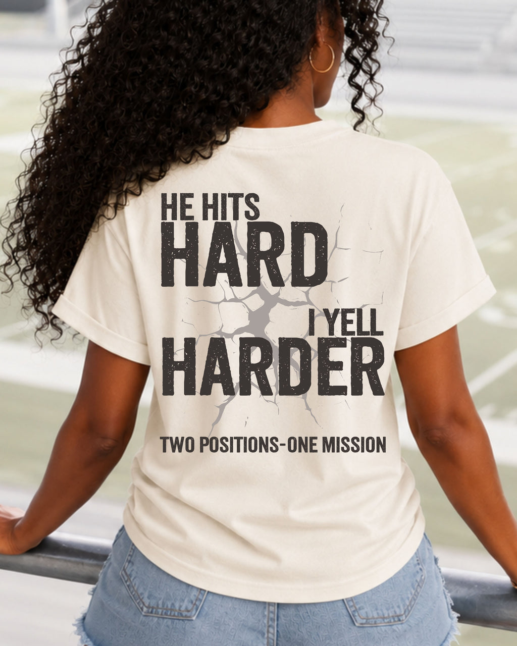 Back view of a woman wearing an ivory Comfort Colors t-shirt in football stadium bleachers featuring the "He Hits Hard I Yell Harder" design with "Two Positions One Mission" on the back.