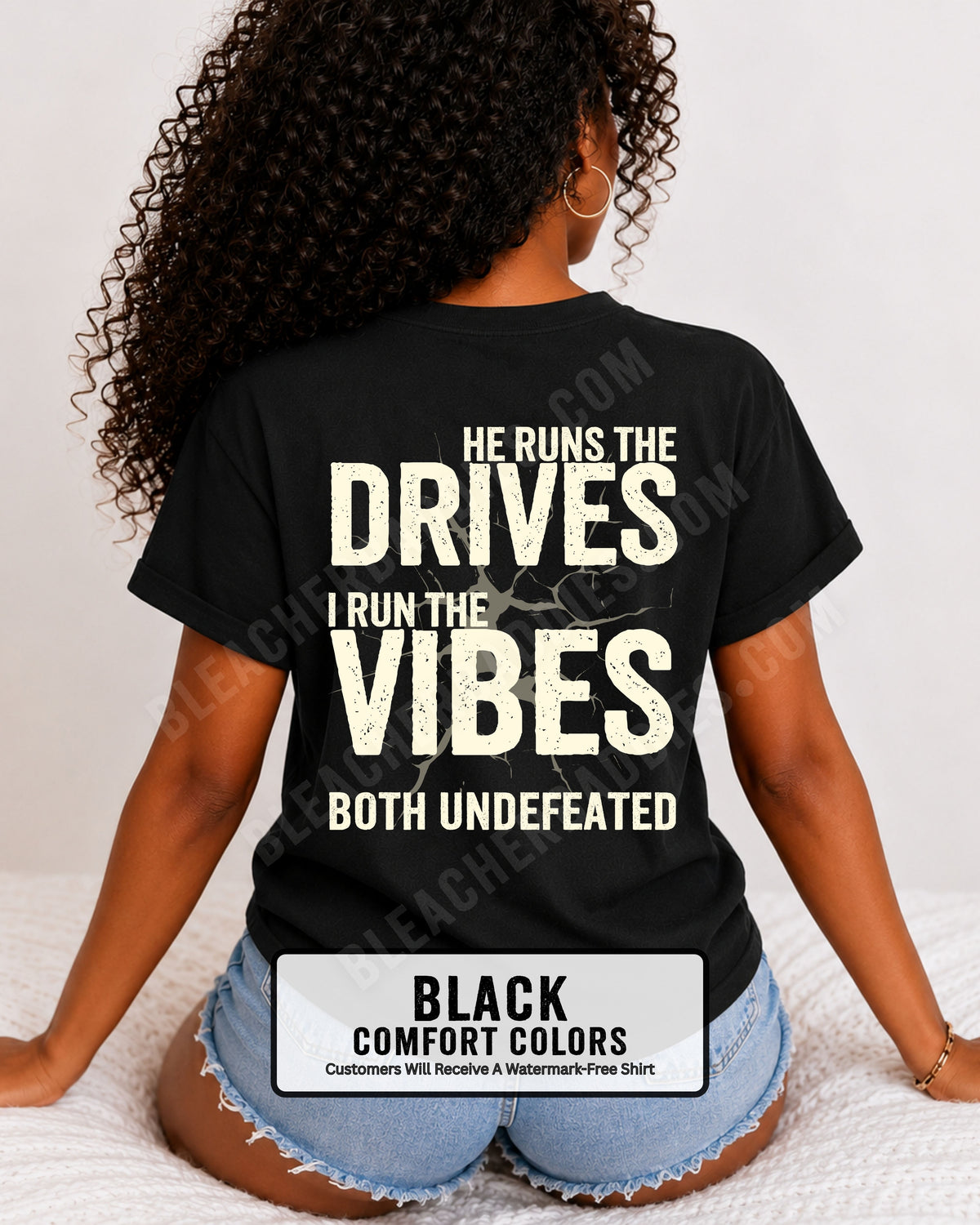 Woman wearing a black Comfort Colors football mom shirt from the back featuring the phrase "He Runs the Drives I Run the Vibes Both Undefeated" in distressed Typography with a cracked background graphic. Running back mom shirt. Quarterback mom shirt Football mom shirt.