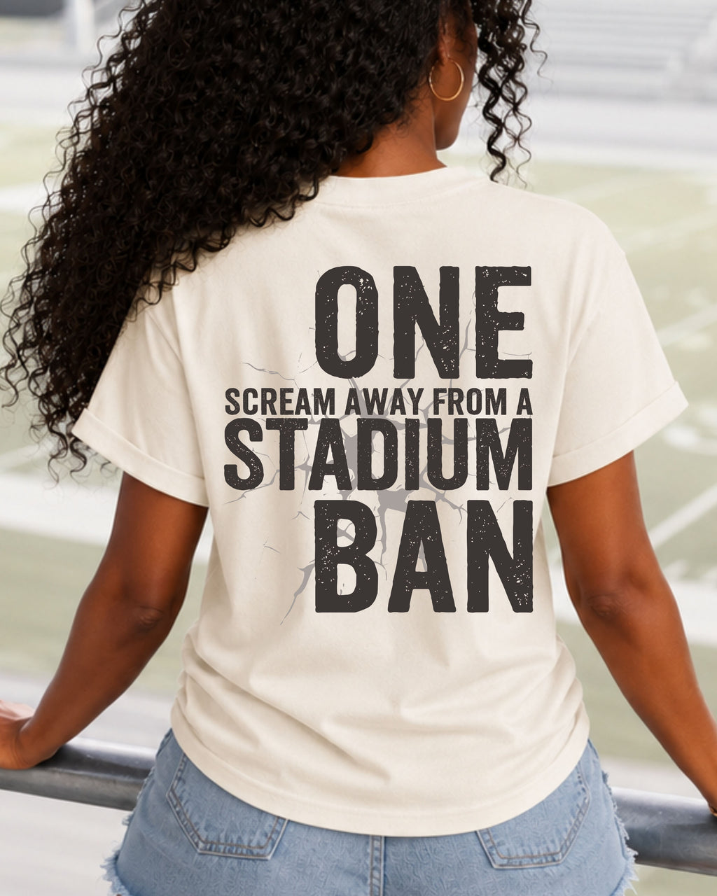 Woman walking through stadium stands wearing an ivory oversized t-shirt with a large distressed black back graphic that reads “One Scream Away From a Stadium Ban” with a cracked texture effect. Football mom shirt. Loud sports mom shirt.