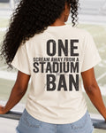 Woman walking through stadium stands wearing an ivory oversized t-shirt with a large distressed black back graphic that reads “One Scream Away From a Stadium Ban” with a cracked texture effect. Football mom shirt. Loud sports mom shirt.