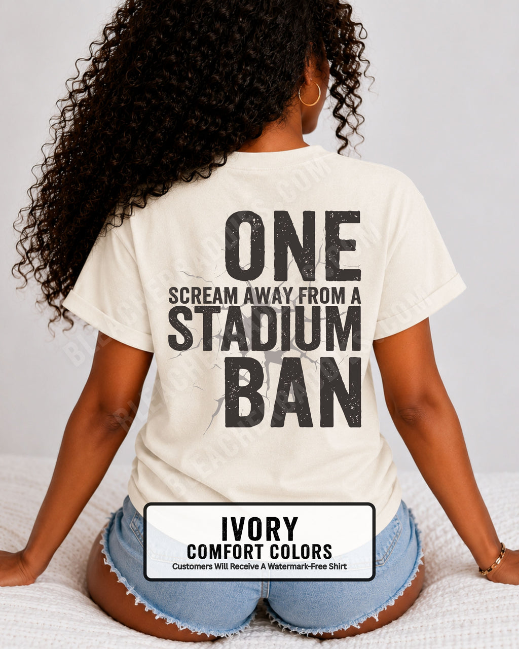 Woman wearing an ivory t-shirt with a large distressed black back graphic that reads “One Scream Away From a Stadium Ban” with a cracked texture effect across the design. Football mom shirt. Loud sports mom shirt.