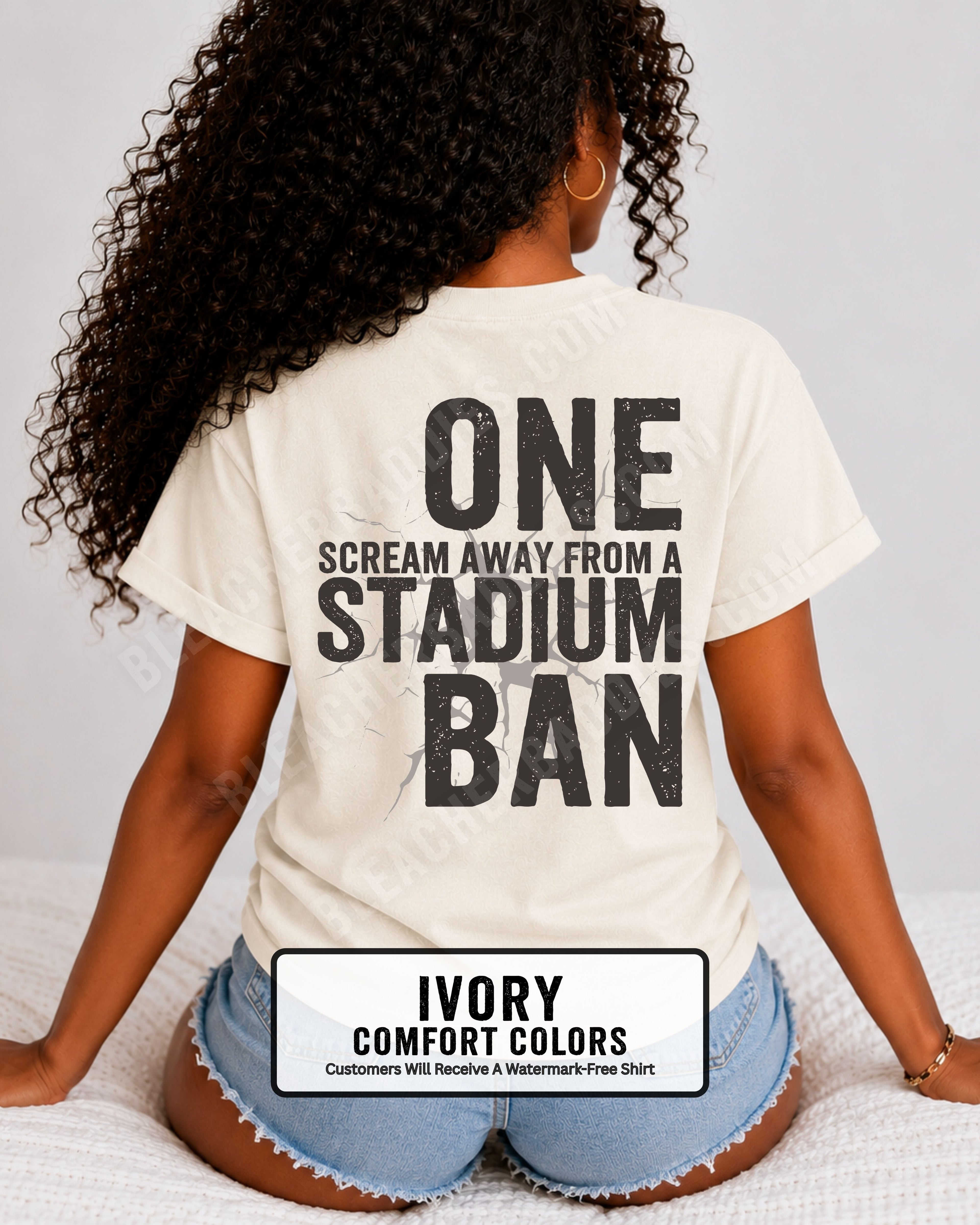 Woman wearing an ivory t-shirt with a large distressed black back graphic that reads “One Scream Away From a Stadium Ban” with a cracked texture effect across the design. Football mom shirt. Loud sports mom shirt.