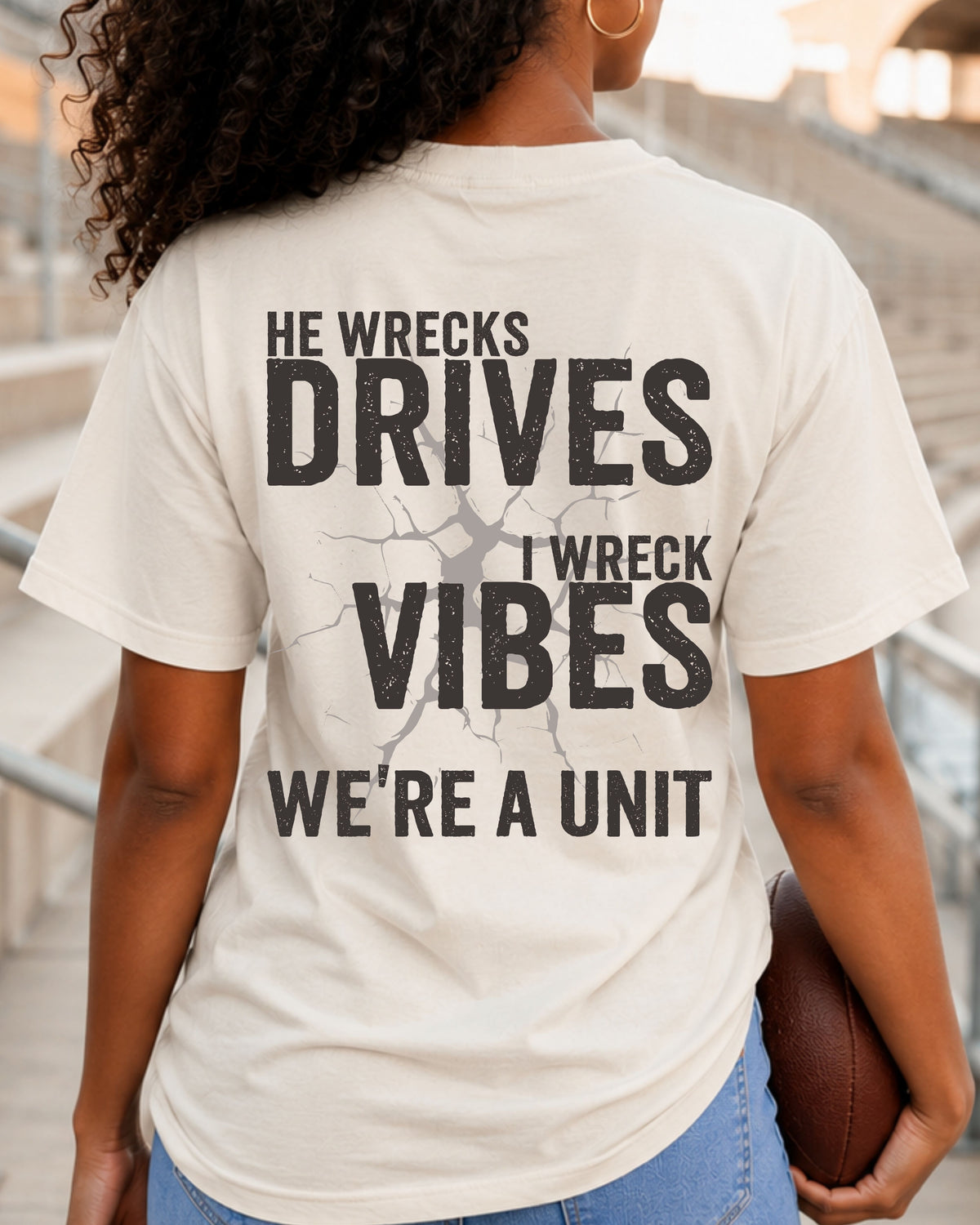 Woman standing in stadium stands wearing an ivory oversized t-shirt with a large distressed black back graphic that reads “He Wrecks Drives I Wreck Vibes We’re a Unit.” Football Mom Shirt. Defensive Lineman End Mom Shirt. Linebacker Mom Shirt. 