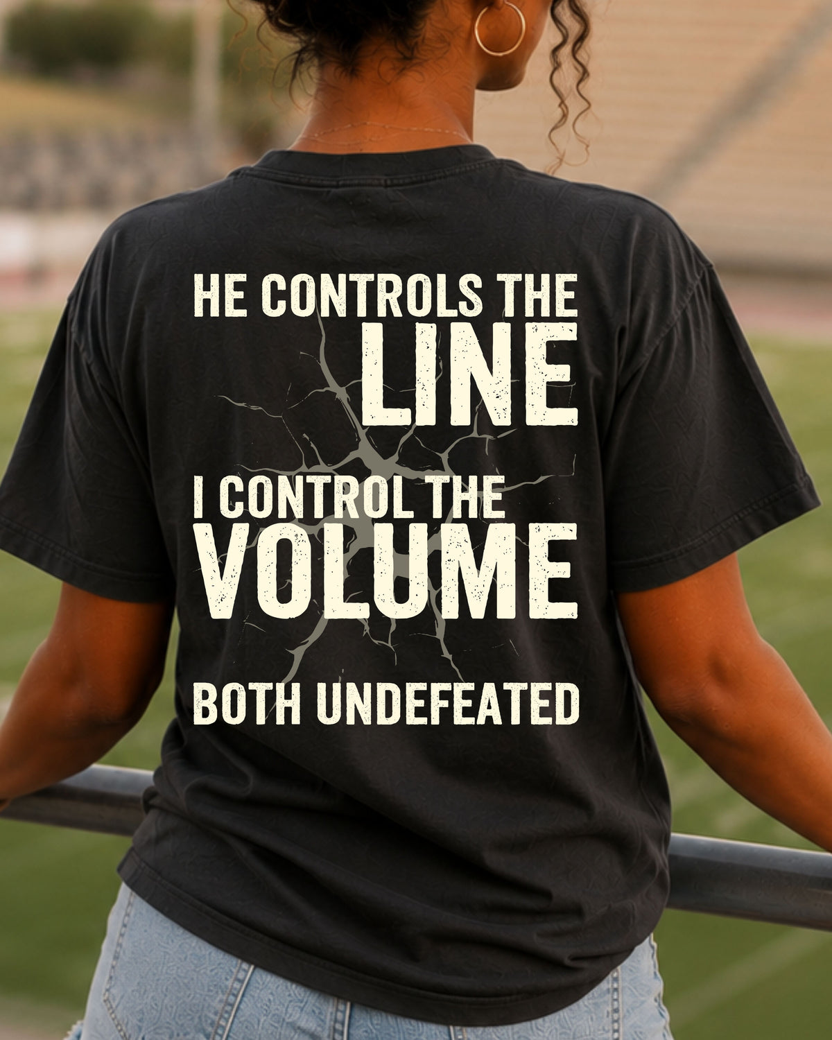 Back view of woman wearing black Comfort Colors football mom game day shirt in stadium featuring “He Controls the Line I Control the Volume Both Undefeated” bold graphic design