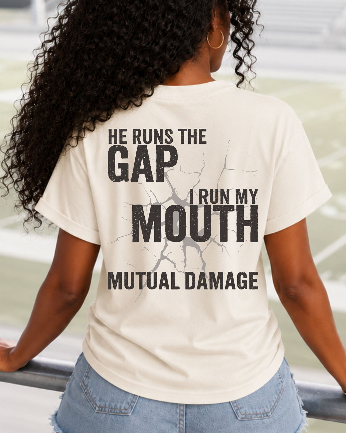 Football mom wearing ivory Comfort Colors game day shirt in stadium with “He Runs the Gap I Run My Mouth Mutual Damage” back print. Running back mom shirt. Quarterback mom shirt. 