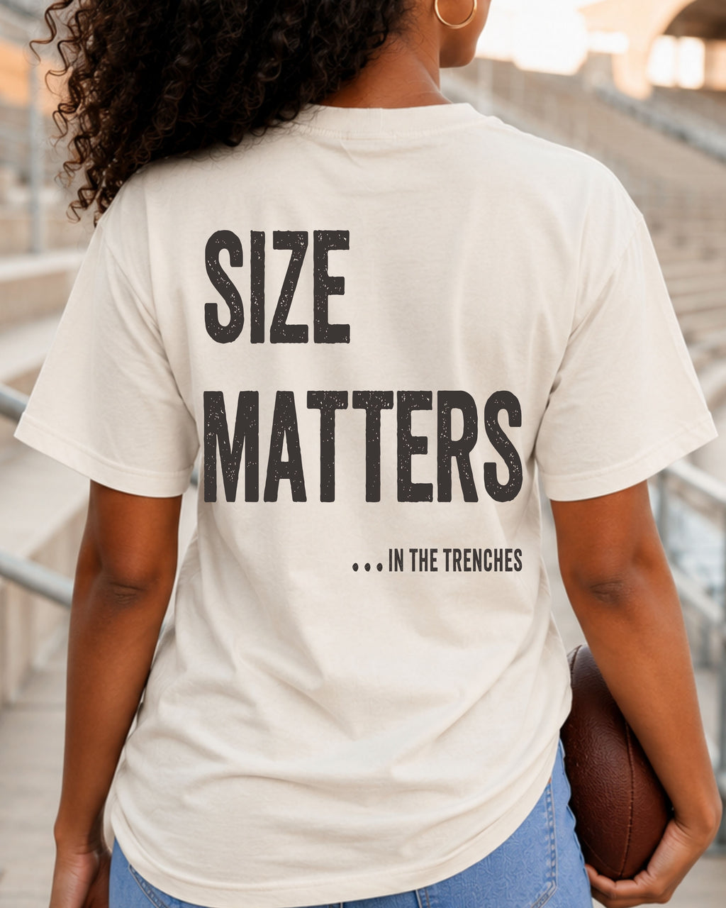 Back view of a woman wearing an ivory Comfort Colors t-shirt with a large distressed "Size Matters... In the Trenches" graphic while holding a football in a stadium. Oversized football fan shirt for offensive line supporters and football season.