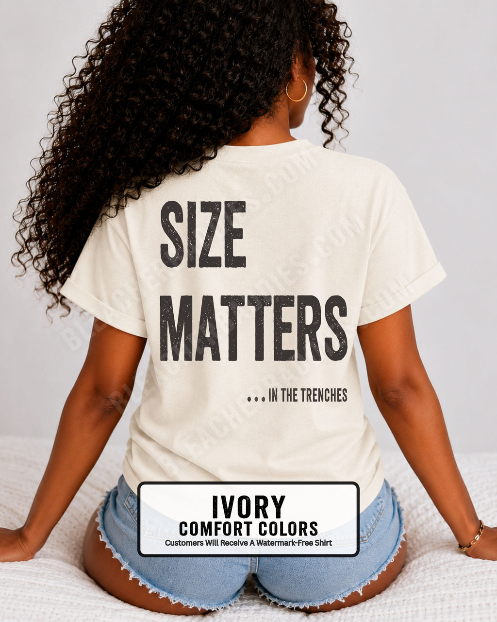 Back view of a woman wearing an ivory Comfort Colors t-shirt with an oversized distressed graphic reading "Size Matters... In the Trenches." Casual football fan shirt styled with denim shorts in a bright lifestyle setting.