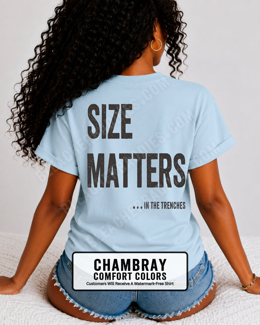 Back view of a woman wearing a CHAMBRAY BLUE Comfort Colors t-shirt with an oversized distressed graphic reading "Size Matters... In the Trenches." Casual football fan shirt styled with denim shorts in a bright lifestyle setting.
