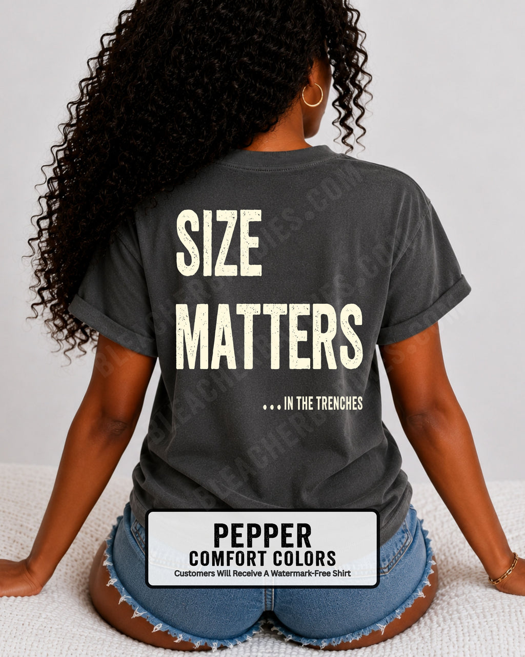 Back view of a woman wearing a PEPPER Comfort Colors t-shirt with an oversized distressed graphic reading "Size Matters... In the Trenches." Casual football fan shirt styled with denim shorts in a bright lifestyle setting.