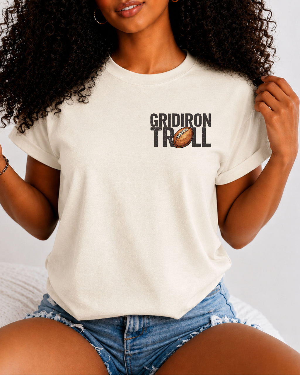 Woman wearing an ivory Comfort Colors t-shirt featuring a small left chest "Gridiron Troll" football graphic with a vintage football replacing the "O." Casual football fan apparel paired with denim shorts in a lifestyle setting.