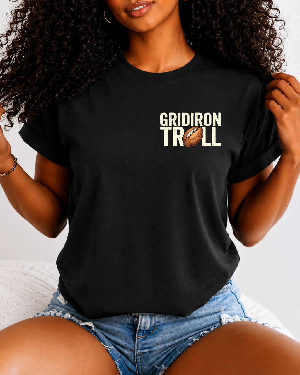 Woman wearing a BLACK Comfort Colors t-shirt featuring a small left chest "Gridiron Troll" football graphic with a vintage football replacing the "O." Casual football fan apparel paired with denim shorts in a lifestyle setting.