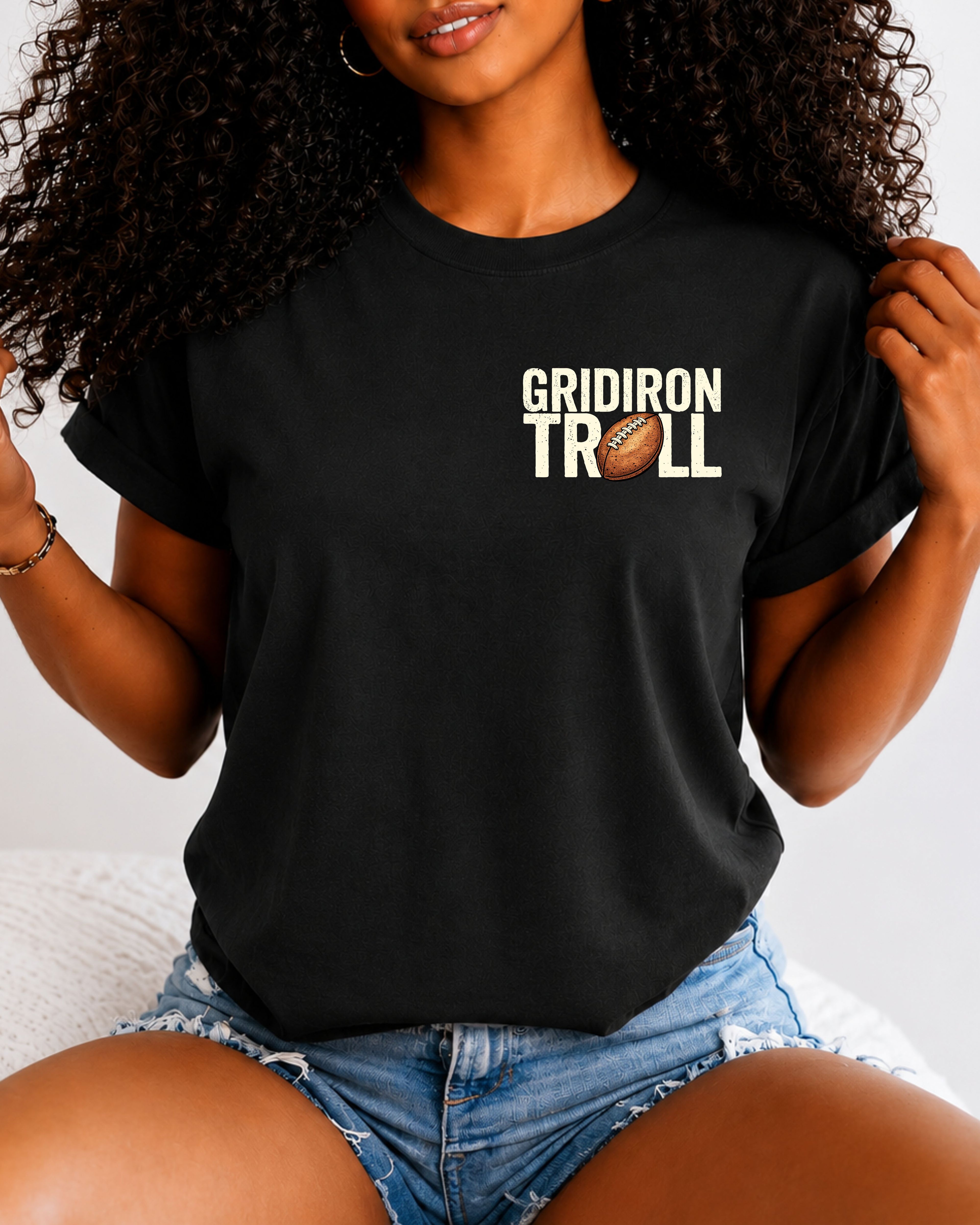 Woman wearing a BLACK Comfort Colors t-shirt featuring a small left chest "Gridiron Troll" football graphic with a vintage football replacing the "O." Casual football fan apparel paired with denim shorts in a lifestyle setting.