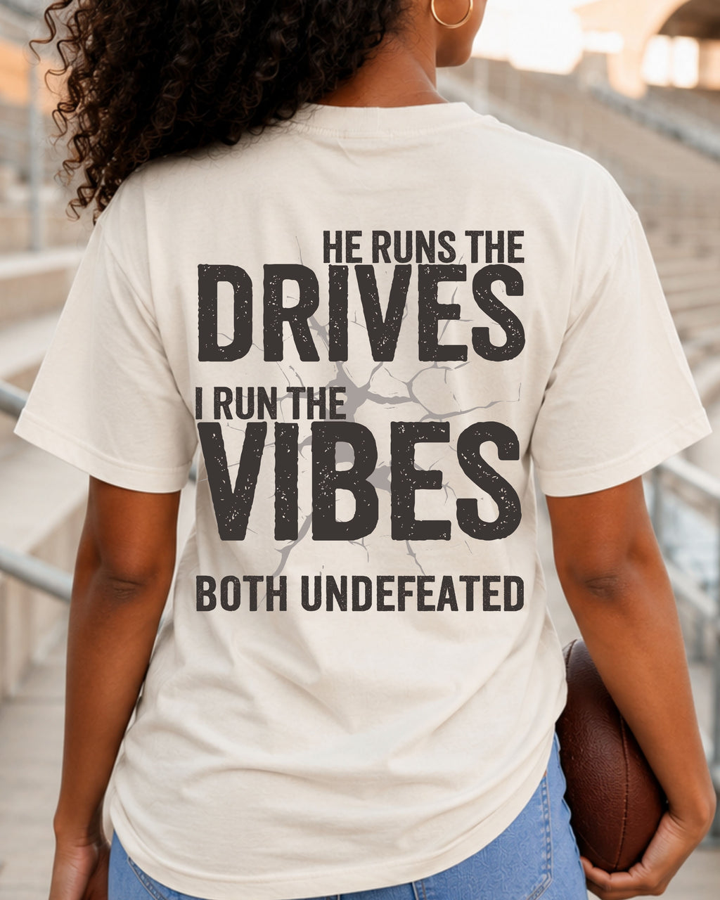 Woman wearing an ivory Comfort Colors football mom shirt from the back in a stadium featuring the phrase "He Runs the Drives I Run the Vibes Both Undefeated" in distressed black lettering with a cracked background design. Running back mom shirt. Quarterback mom shirt. Football mom shirt.