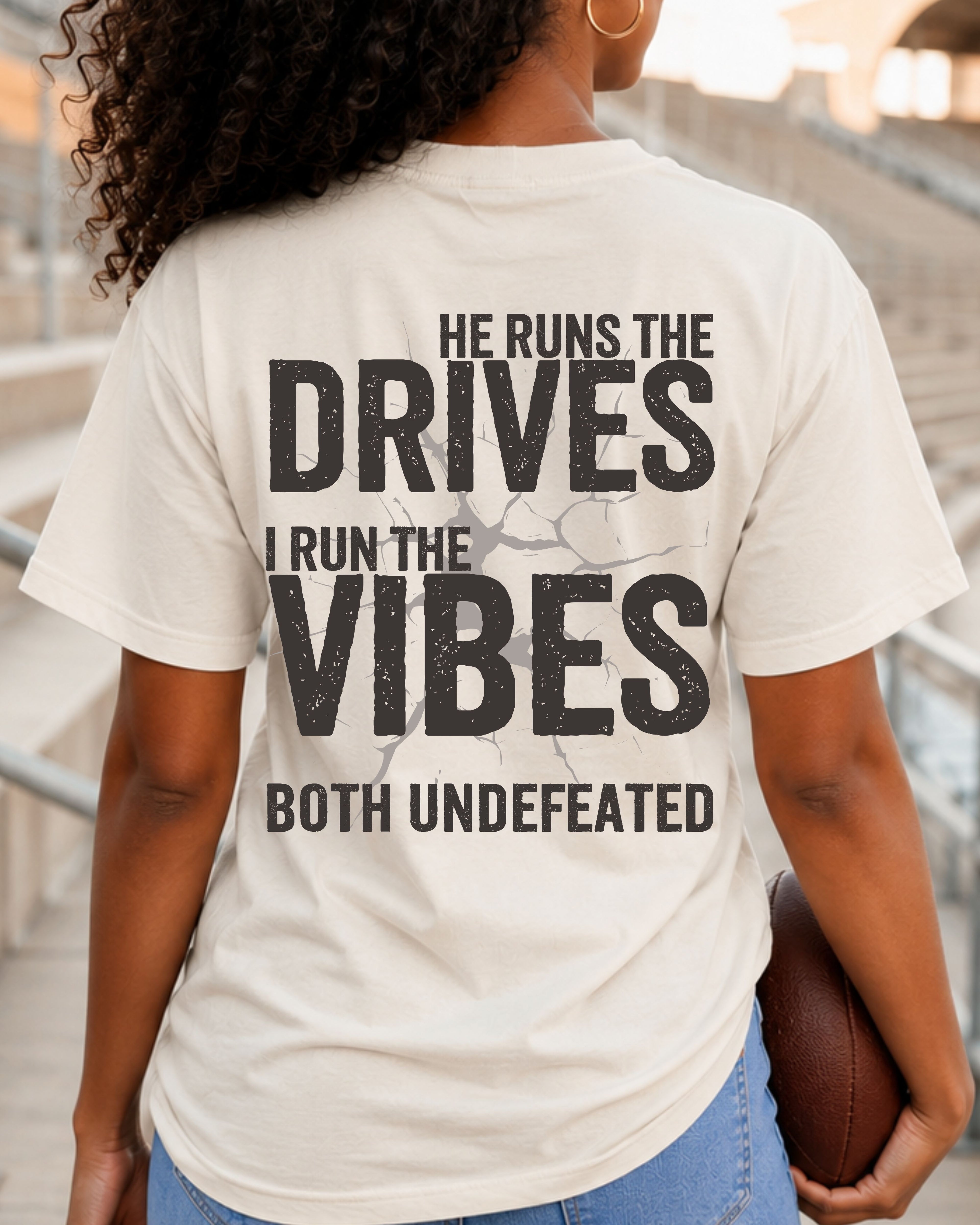 Woman wearing an ivory Comfort Colors football mom shirt from the back in a stadium featuring the phrase "He Runs the Drives I Run the Vibes Both Undefeated" in distressed black lettering with a cracked background design. Running back mom shirt. Quarterback mom shirt. Football mom shirt.