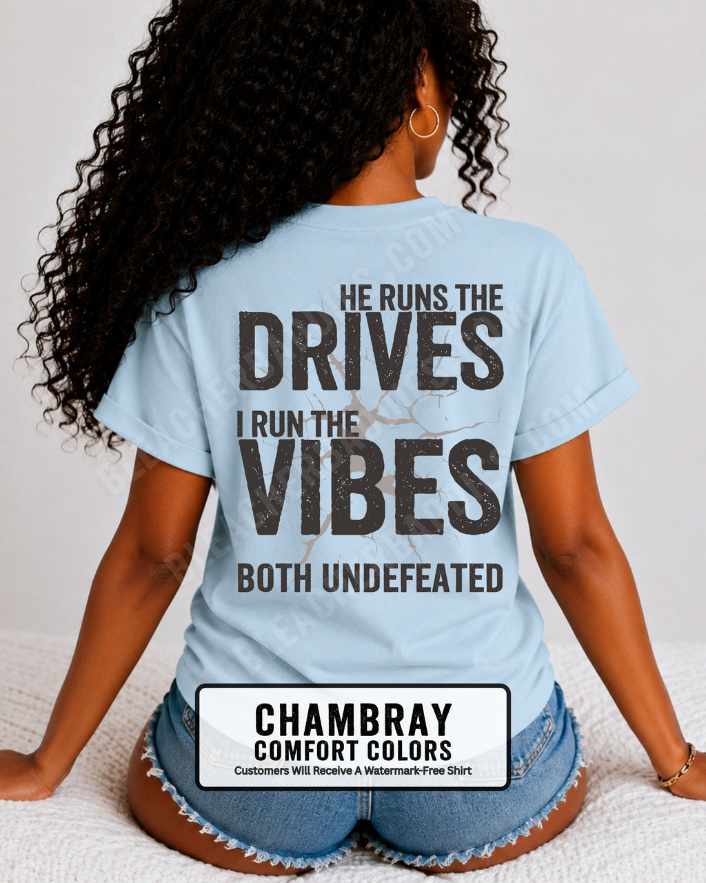 Woman wearing a chambray blue Comfort Colors football mom shirt from the back featuring the phrase "He Runs the Drives I Run the Vibes Both Undefeated" in distressed Typography with a cracked background graphic. Running back mom shirt. Quarterback mom shirt Football mom shirt.