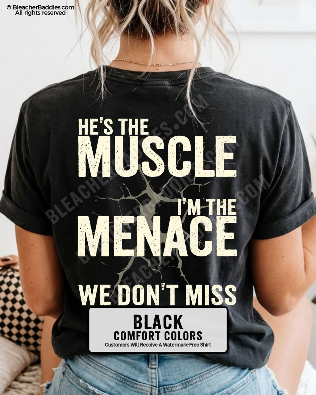 Woman wearing BLACK Comfort Colors football mom game day shirt with back design that reads “He’s the muscle I’m the menace we don’t miss” in distressed CREAM text with cracked graphic and chambray color label shown