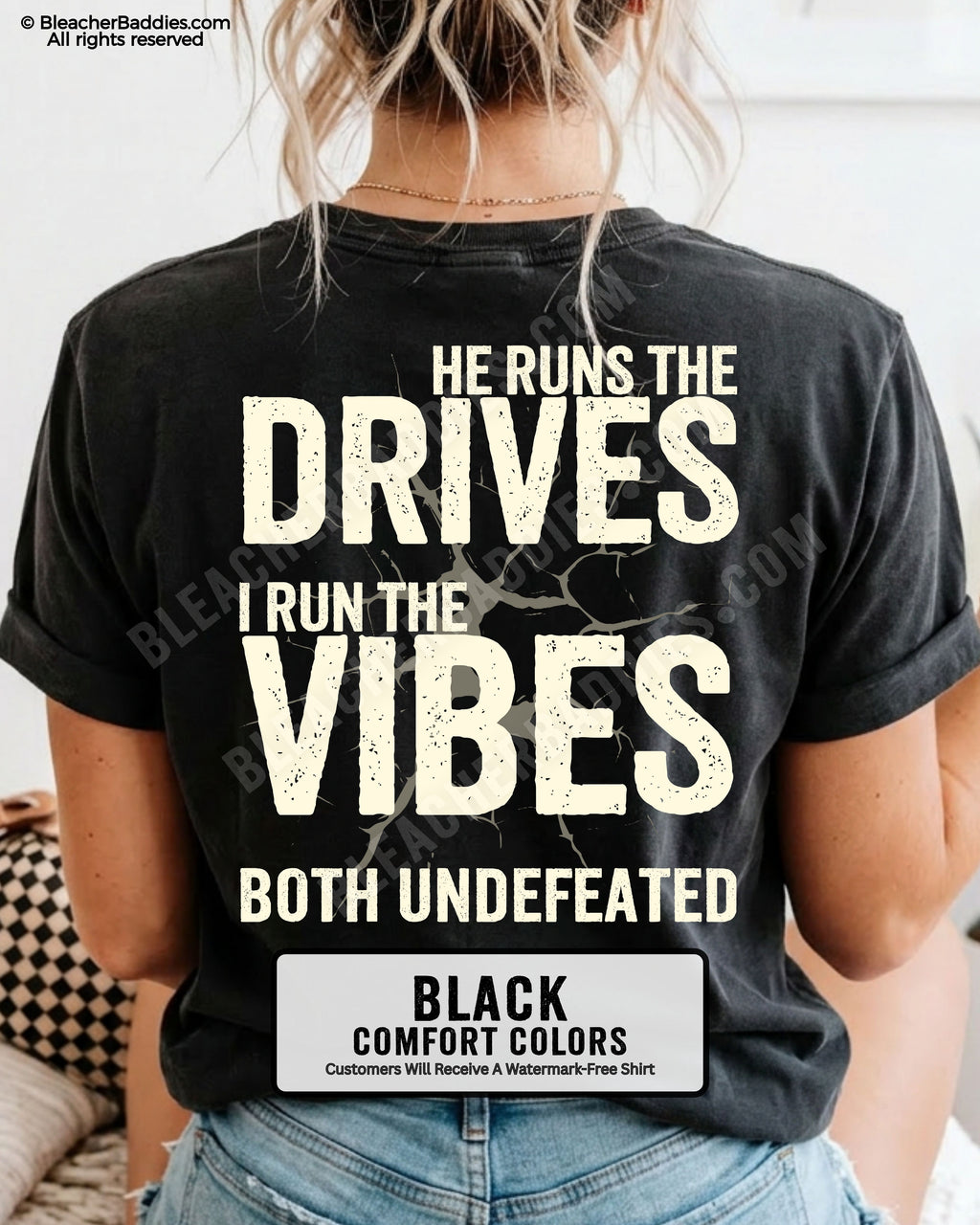 Woman wearing a black Comfort Colors football mom shirt from the back featuring the phrase "He Runs the Drives I Run the Vibes Both Undefeated" in distressed Typography with a cracked background graphic.