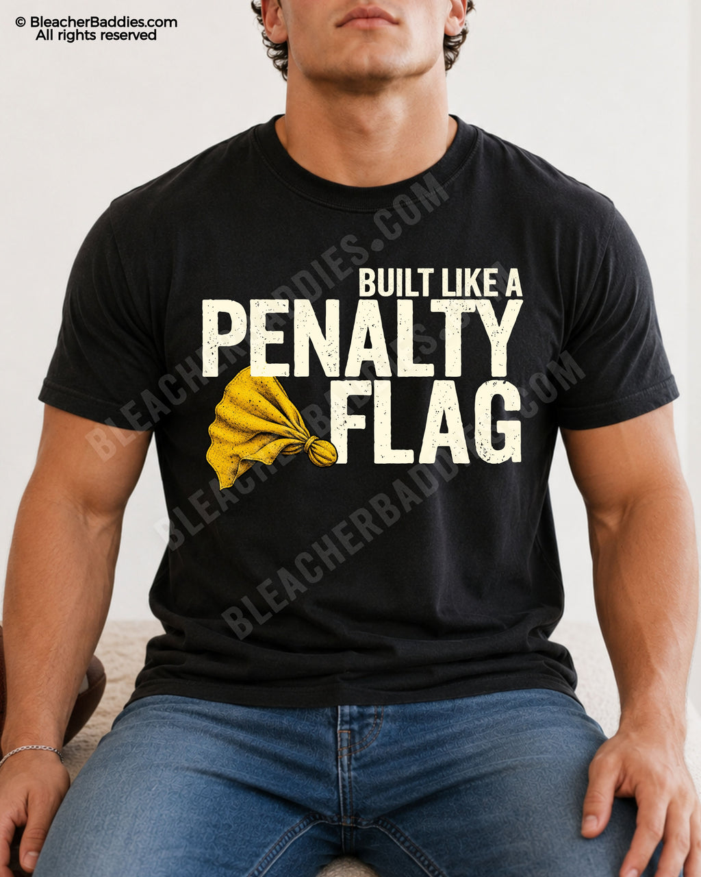 Muscular young male football player model sitting indoors wearing a black Comfort Colors style shirt with distressed “Built Like A Penalty Flag” graphic and yellow penalty flag illustration paired with blue jeans.
