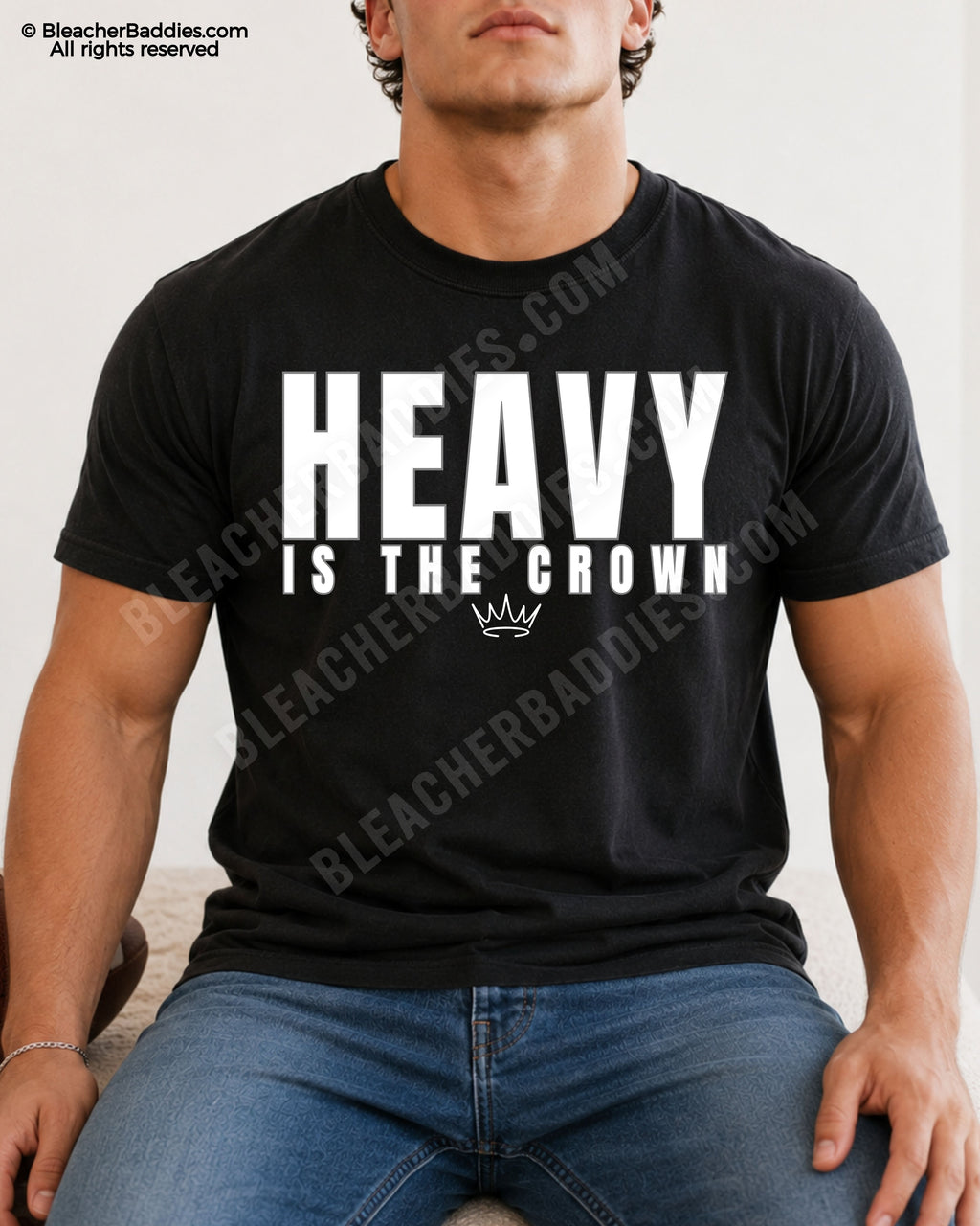 Young male athlete wearing a black “HEAVY IS THE CROWN” motivational football graphic t-shirt while sitting indoors in blue jeans with a relaxed athletic pose.