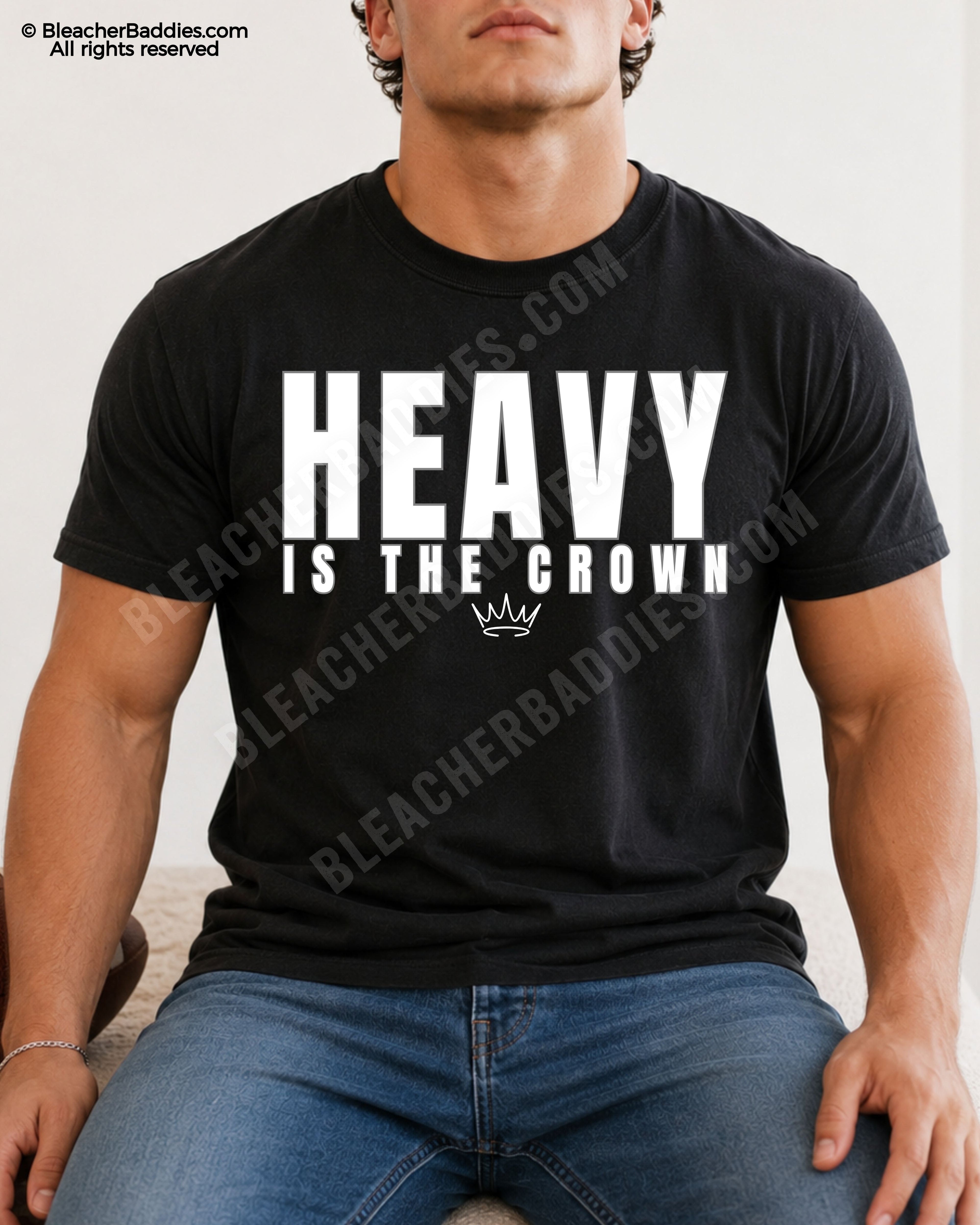 Young male athlete wearing a black “HEAVY IS THE CROWN” motivational football graphic t-shirt while sitting indoors in blue jeans with a relaxed athletic pose.