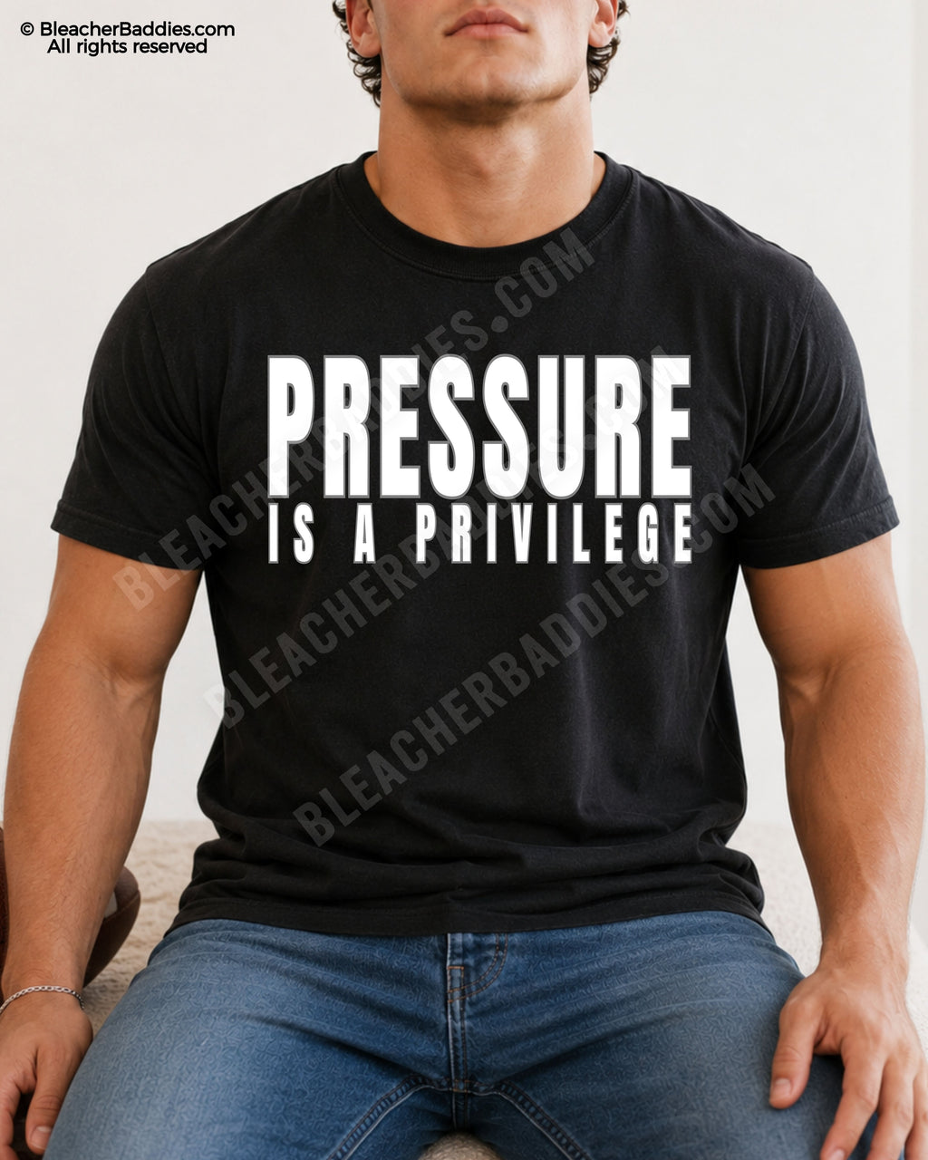 Young male athlete wearing a BLACK motivational football graphic t-shirt with bold WHITE “PRESSURE IS A PRIVILEGE” lettering while sitting indoors in jeans.