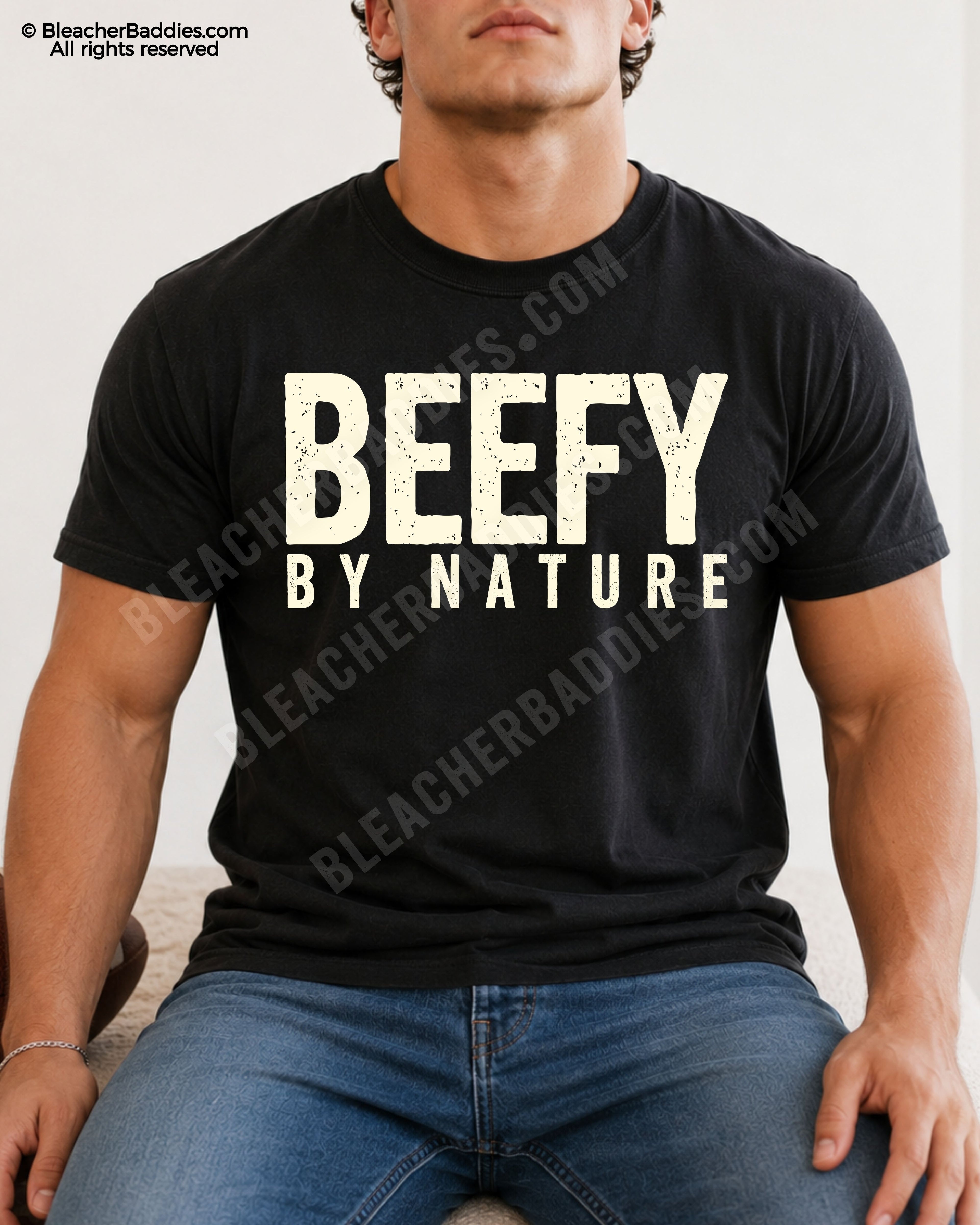 Male football player model wearing a black Comfort Colors “Beefy By Nature” football shirt with distressed cream typography while seated indoors in jeans.