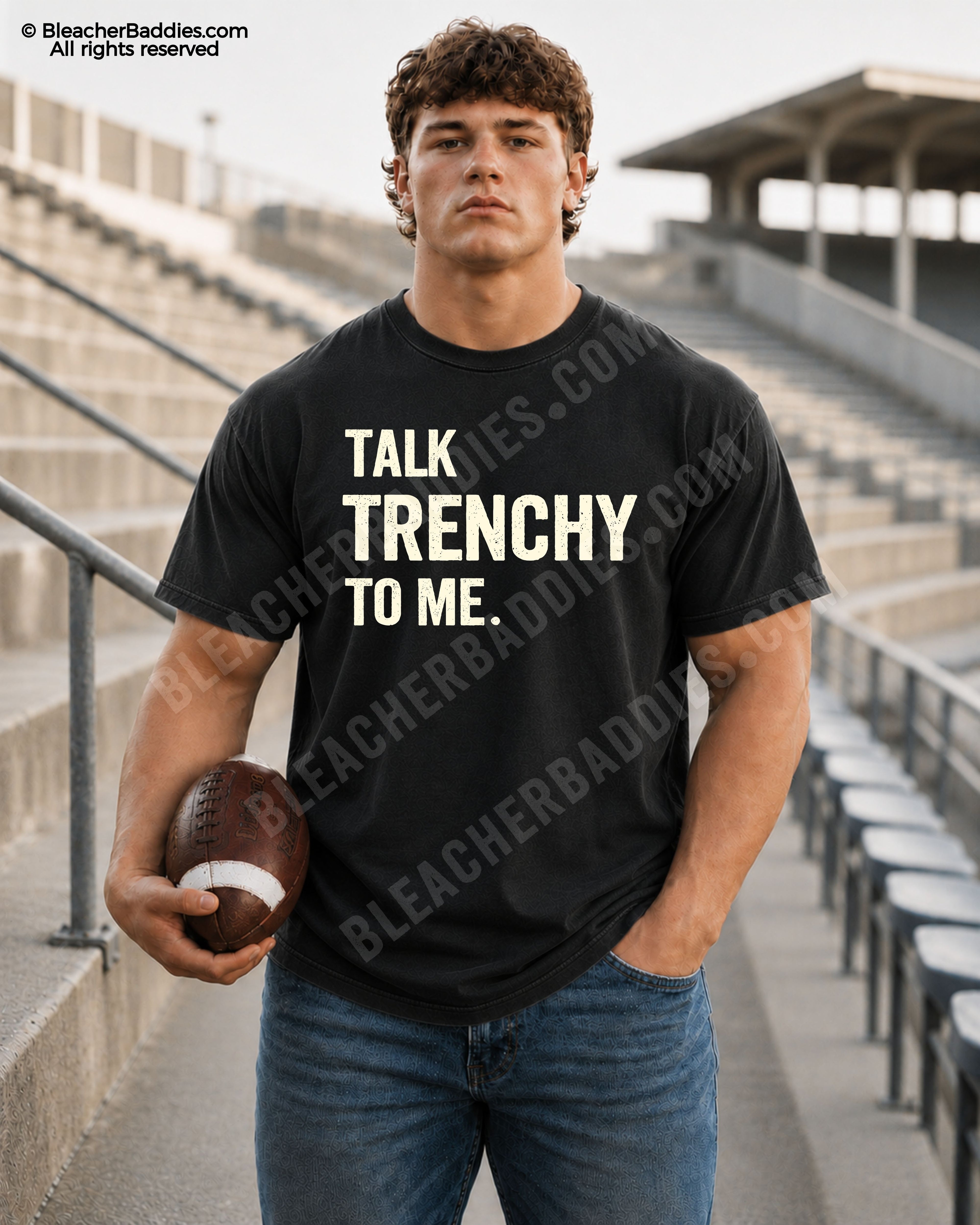 Athletic male model wearing a black Comfort Colors 1717 t-shirt with "Talk Trenchy to Me" graphic, distressed blue jeans, and holding a football in empty stadium bleachers.