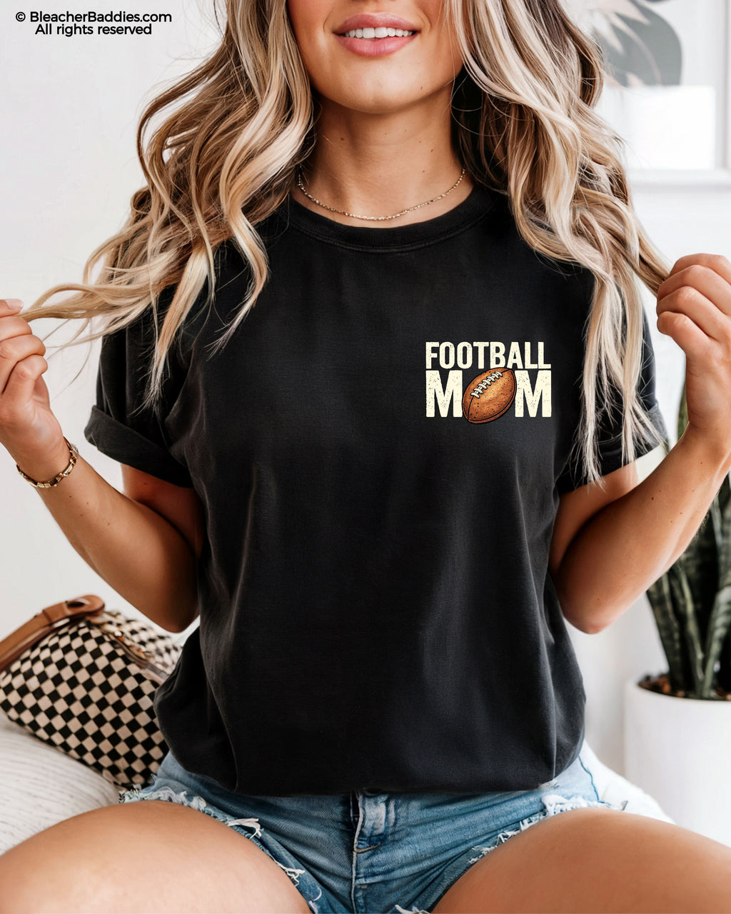 Woman wearing a black t-shirt with a small left chest graphic that reads “Football Mom” with a brown football replacing the “O” in mom, styled with a relaxed fit and paired with denim shorts.