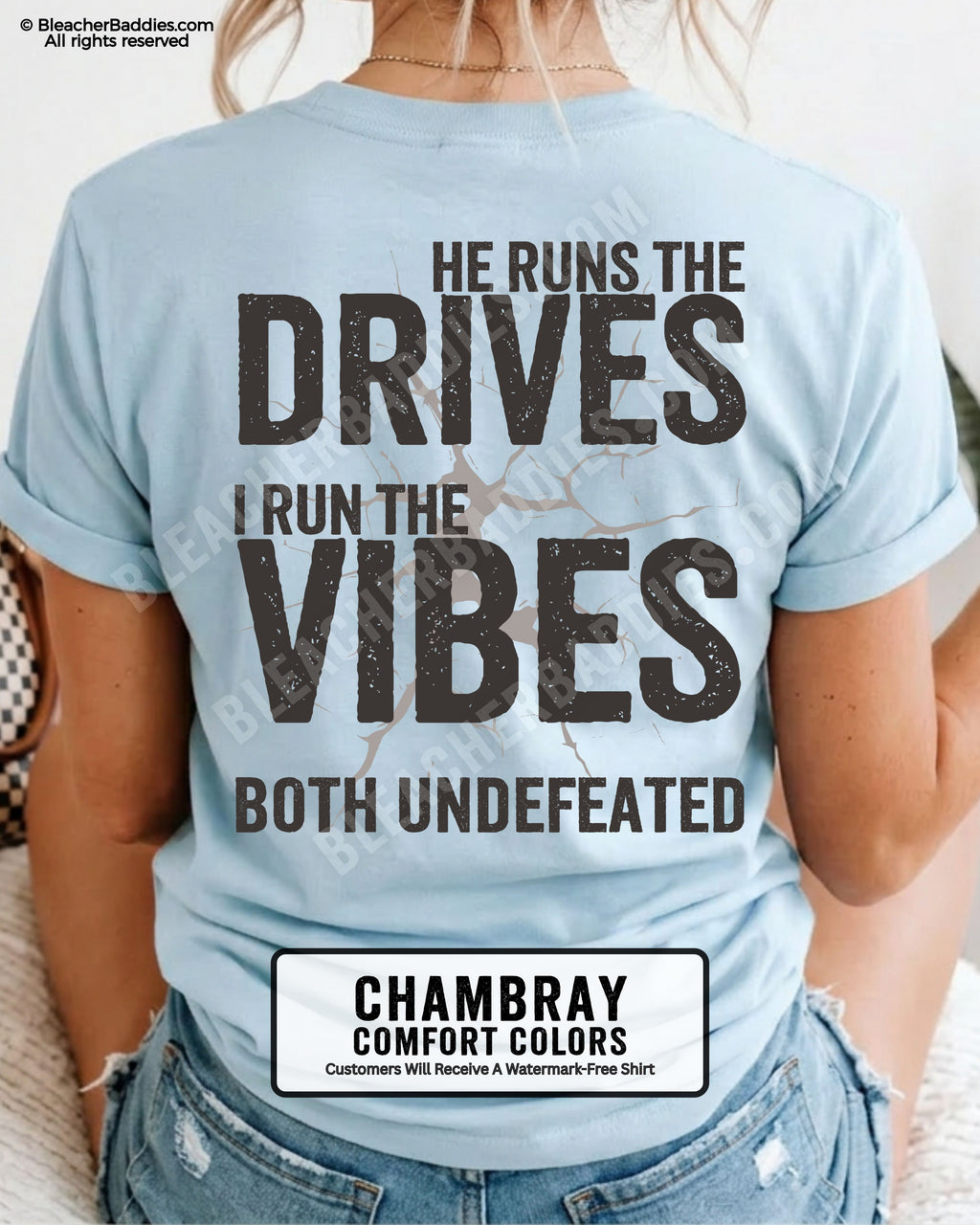 Woman wearing a chambray blue Comfort Colors football mom shirt from the back featuring the phrase "He Runs the Drives I Run the Vibes Both Undefeated" in distressed Typography with a cracked background graphic.