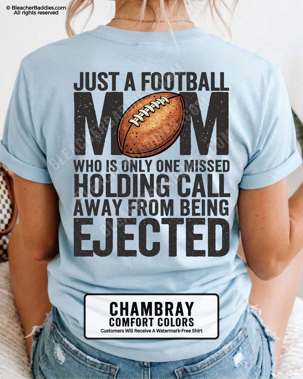 Back view of a woman wearing a chambray Comfort Colors football mom shirt, displaying bold distressed text that reads “Just a football mom who is only one missed holding call away from being ejected,” with a football graphic on the back.