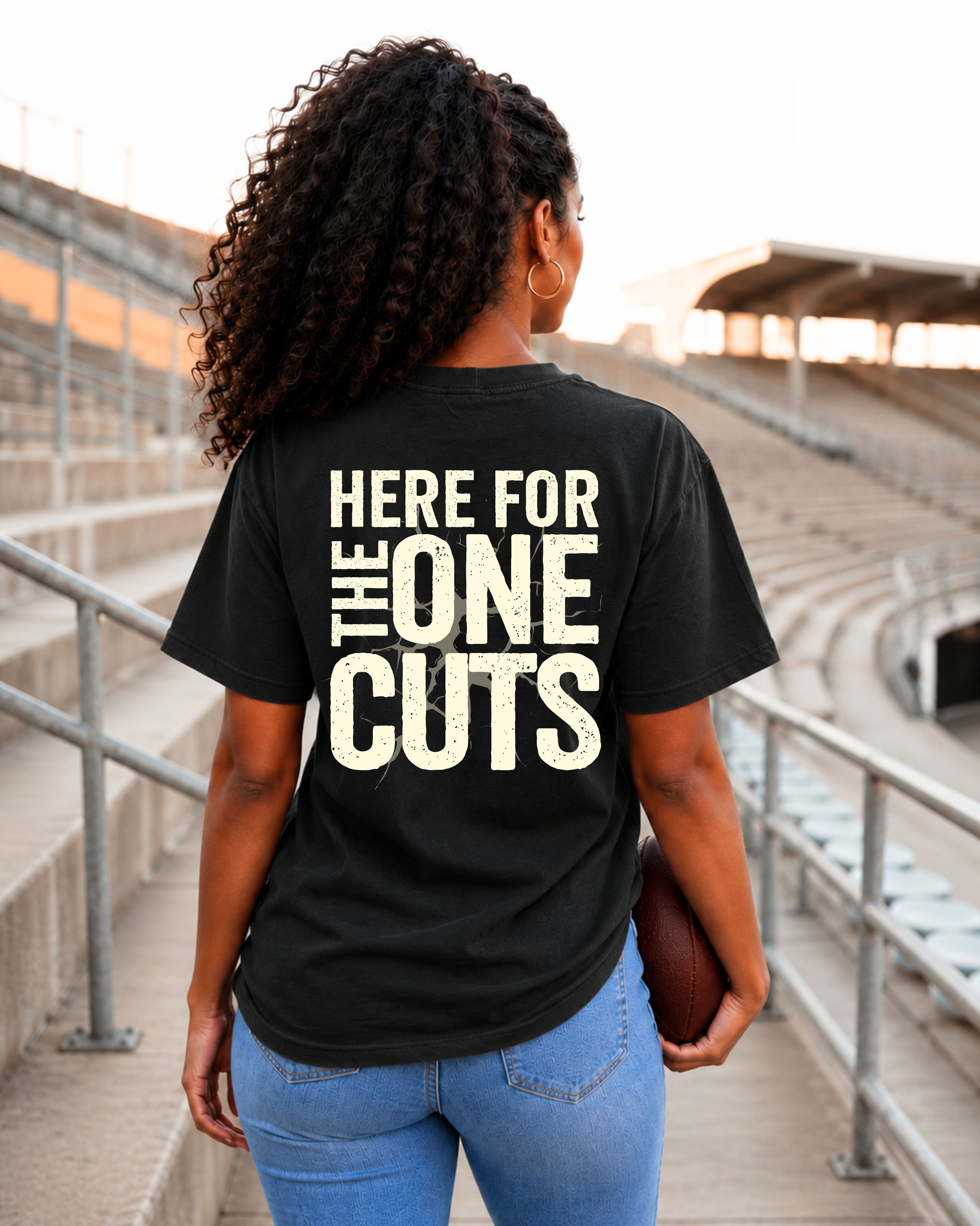 Model wearing a black Comfort Colors t-shirt in a football stadium featuring the distressed black phrase "HERE FOR THE ONE CUTS" printed on the back. Oversized unisex graphic tee for football moms, football fans, game days, tailgates, Friday night lights, and stadium style.