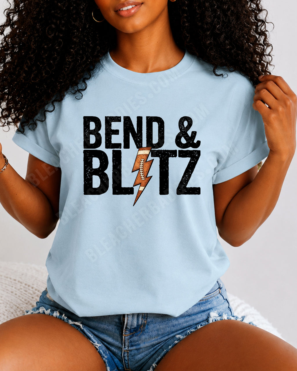 Close-up lifestyle photo of a football mom wearing a chambray Comfort Colors t-shirt with the distressed black graphic "BEND & BLITZ" and a football lightning bolt design between the words. 