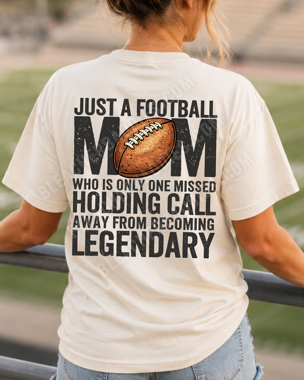 A woman stands on a stadium railing at sunset, viewed from behind, wearing an ivory comfort colors t-shirt with a large black and brown football graphic and text that reads 'JUST A FOOTBALL MOM WHO IS ONLY ONE MISSED HOLDING CALL AWAY FROM BECOMING LEGENDARY'.