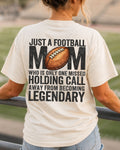 A woman stands on a stadium railing at sunset, viewed from behind, wearing an ivory comfort colors t-shirt with a large black and brown football graphic and text that reads 'JUST A FOOTBALL MOM WHO IS ONLY ONE MISSED HOLDING CALL AWAY FROM BECOMING LEGENDARY'.