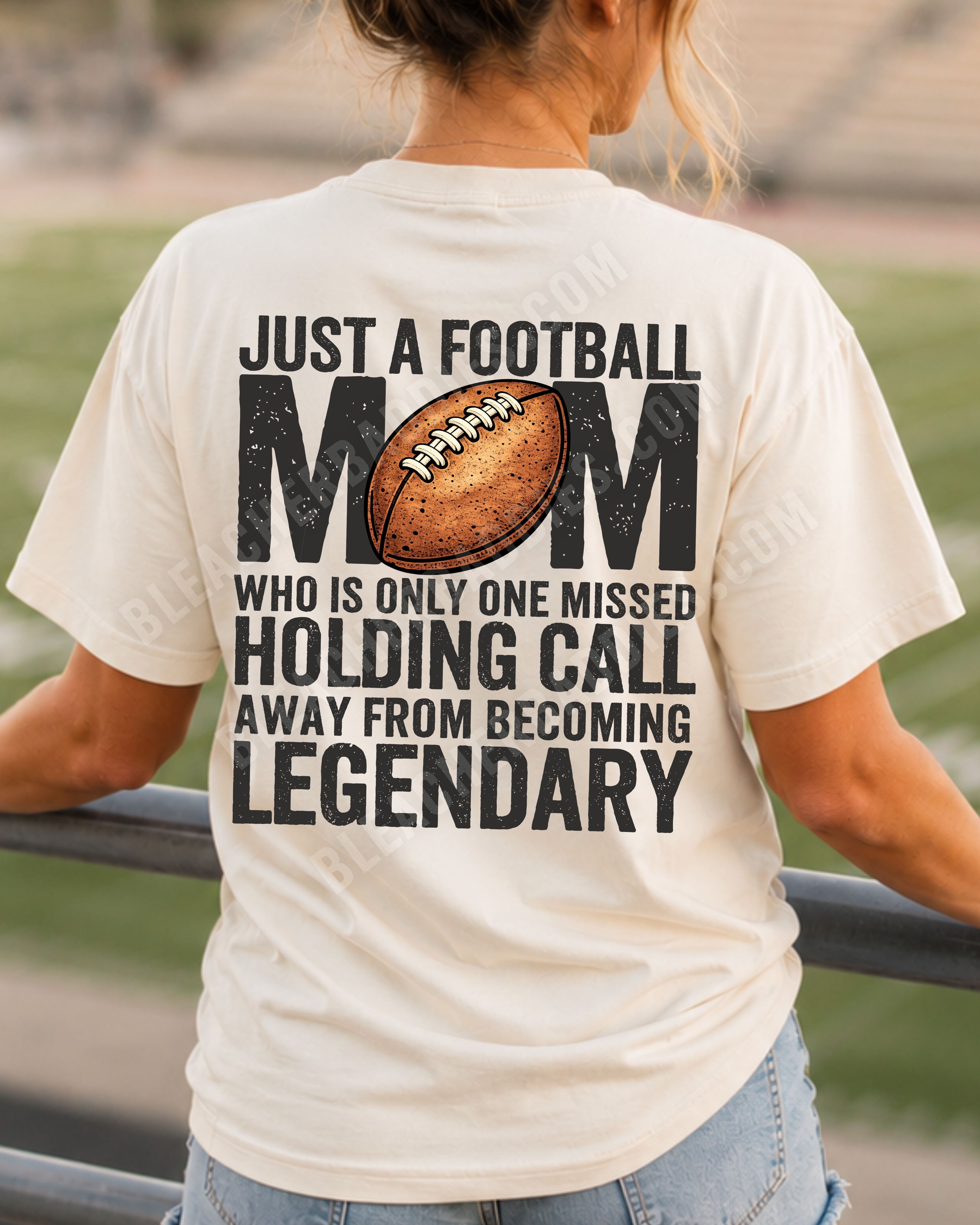 A woman stands on a stadium railing at sunset, viewed from behind, wearing an ivory comfort colors t-shirt with a large black and brown football graphic and text that reads 'JUST A FOOTBALL MOM WHO IS ONLY ONE MISSED HOLDING CALL AWAY FROM BECOMING LEGENDARY'.