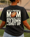 A woman viewed from behind wearing a black graphic t-shirt with a football-themed design. The shirt features the text 'JUST A FOOTBALL MOM WHO DOESN'T EVER SIT DOWN OR SHUT UP' in distressed white lettering, with a brown football graphic replacing the letter 'O'.