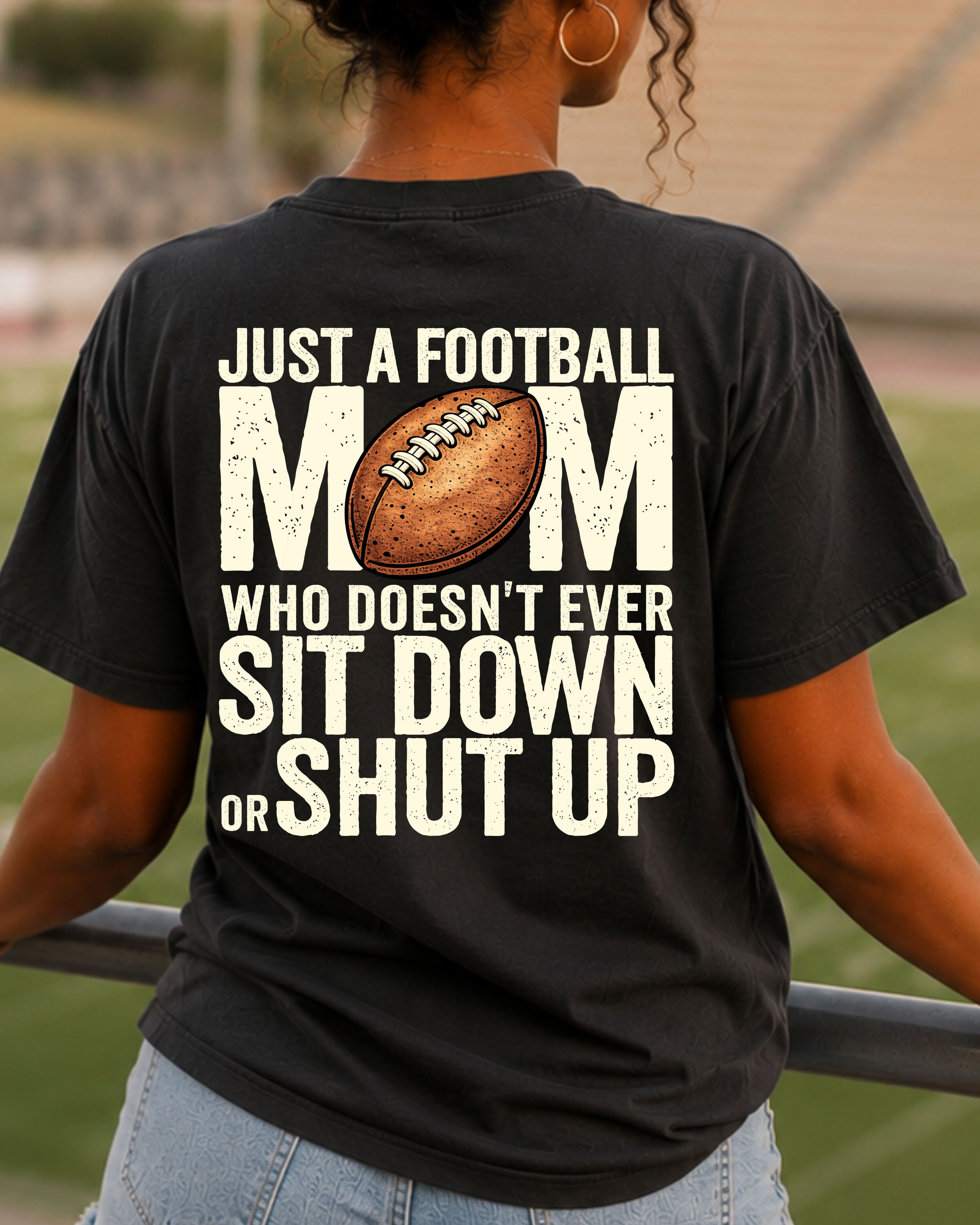 A woman viewed from behind wearing a black graphic t-shirt with a football-themed design. The shirt features the text 'JUST A FOOTBALL MOM WHO DOESN'T EVER SIT DOWN OR SHUT UP' in distressed white lettering, with a brown football graphic replacing the letter 'O'.