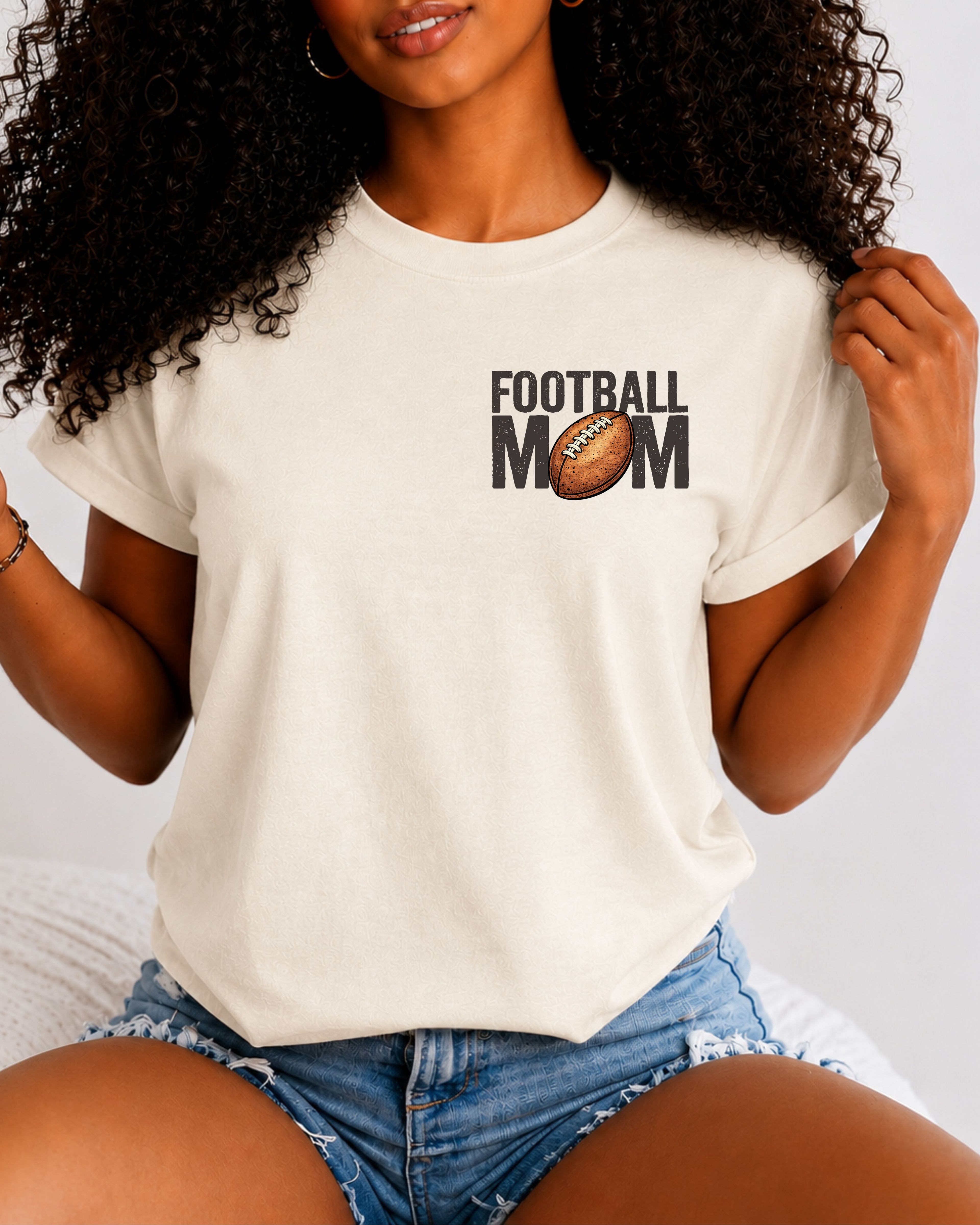 Woman wearing an ivory t-shirt with a small left chest graphic that reads “Football Mom” with a brown football replacing the “O” in mom, styled with a relaxed fit and paired with denim shorts.
