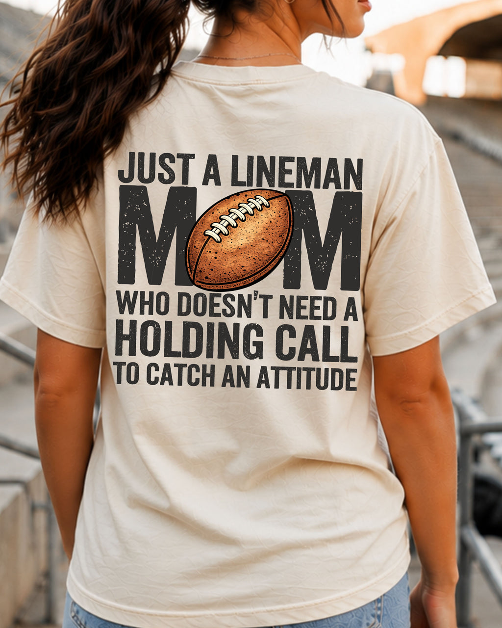 Woman wearing ivory Comfort Colors lineman mom football game day shirt with back print that says “Just a lineman mom who doesn’t need a holding call to catch an attitude”