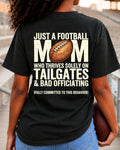 Football mom game day shirt worn by a model in a stadium, shown on a black Comfort Colors tee with a cream graphic that says, “Just a football mom who thrives solely on tailgates & bad officiating (fully committed to this behavior).”