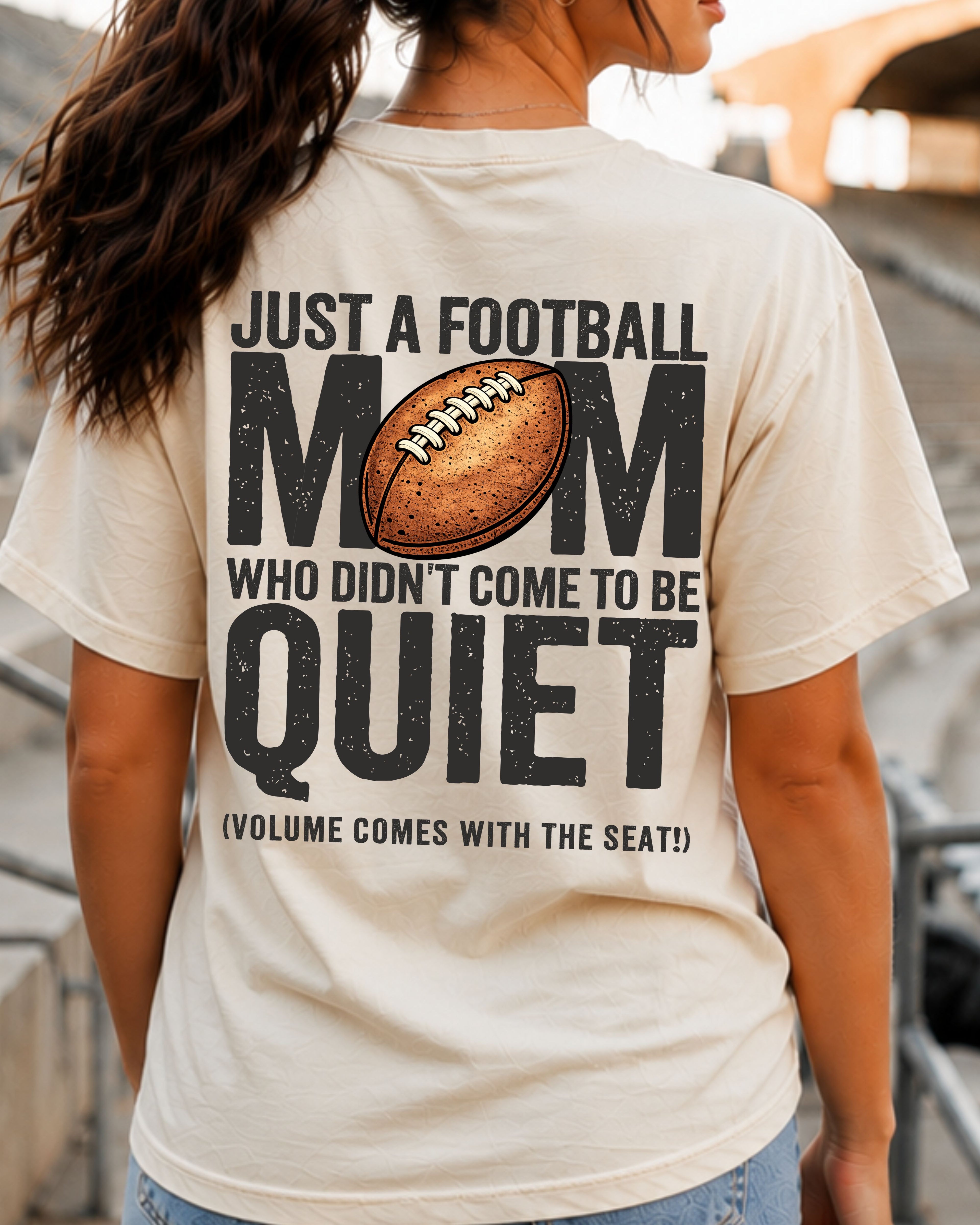 Football mom game day shirt worn in stadium stands on an ivory Comfort Colors tee with back design reading “Just a football mom who didn’t come to be quiet volume comes with the seat”