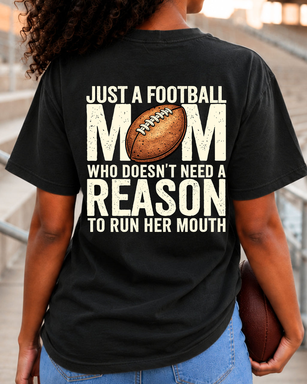 Woman wearing black Comfort Colors t-shirt in stadium bleachers featuring large back print that says “Just a Football Mom Who Doesn’t Need a Reason to Run Her Mouth” with football graphic