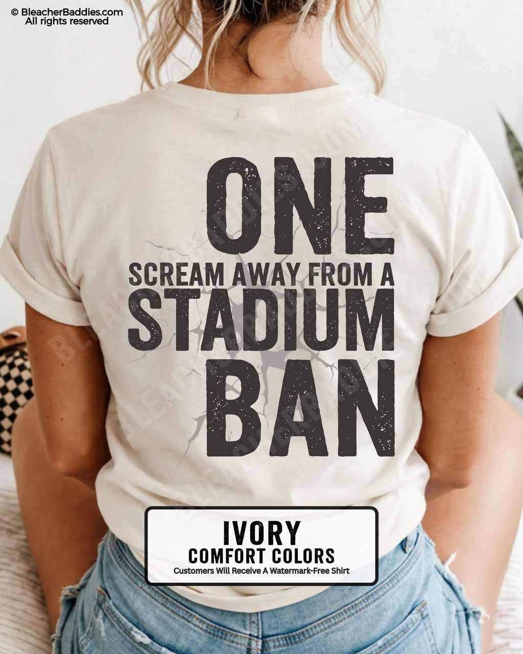 Woman wearing an ivory t-shirt with a large distressed black back graphic that reads “One Scream Away From a Stadium Ban” with a cracked texture effect across the design.