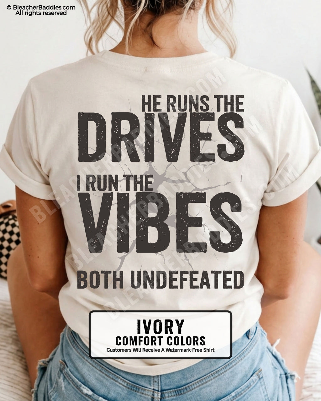 Woman wearing an ivory Comfort Colors football mom shirt from the back featuring the phrase "He Runs the Drives I Run the Vibes Both Undefeated" in distressed Typography with a cracked background graphic.