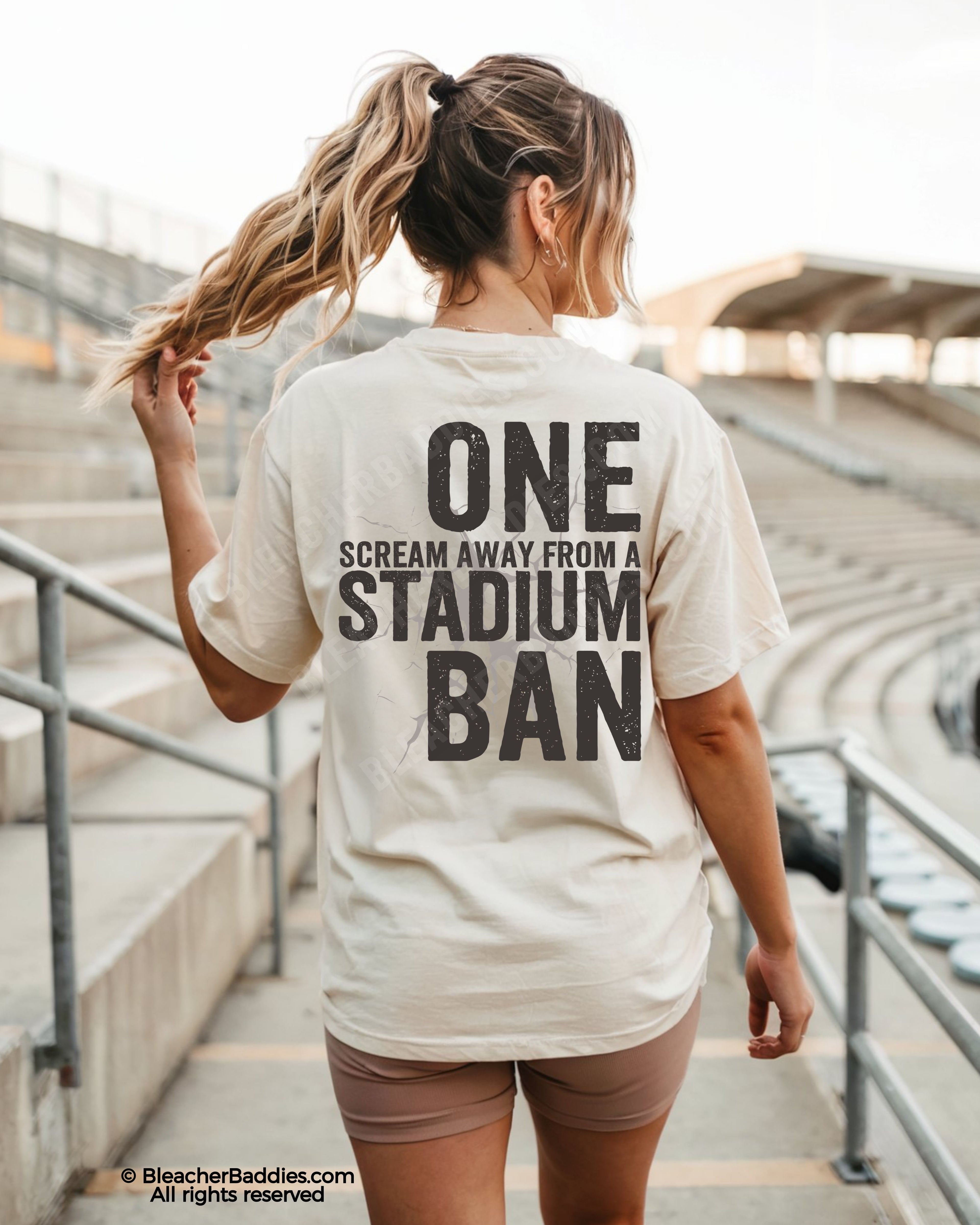 Woman walking through stadium stands wearing an ivory oversized t-shirt with a large distressed black back graphic that reads “One Scream Away From a Stadium Ban” with a cracked texture effect.