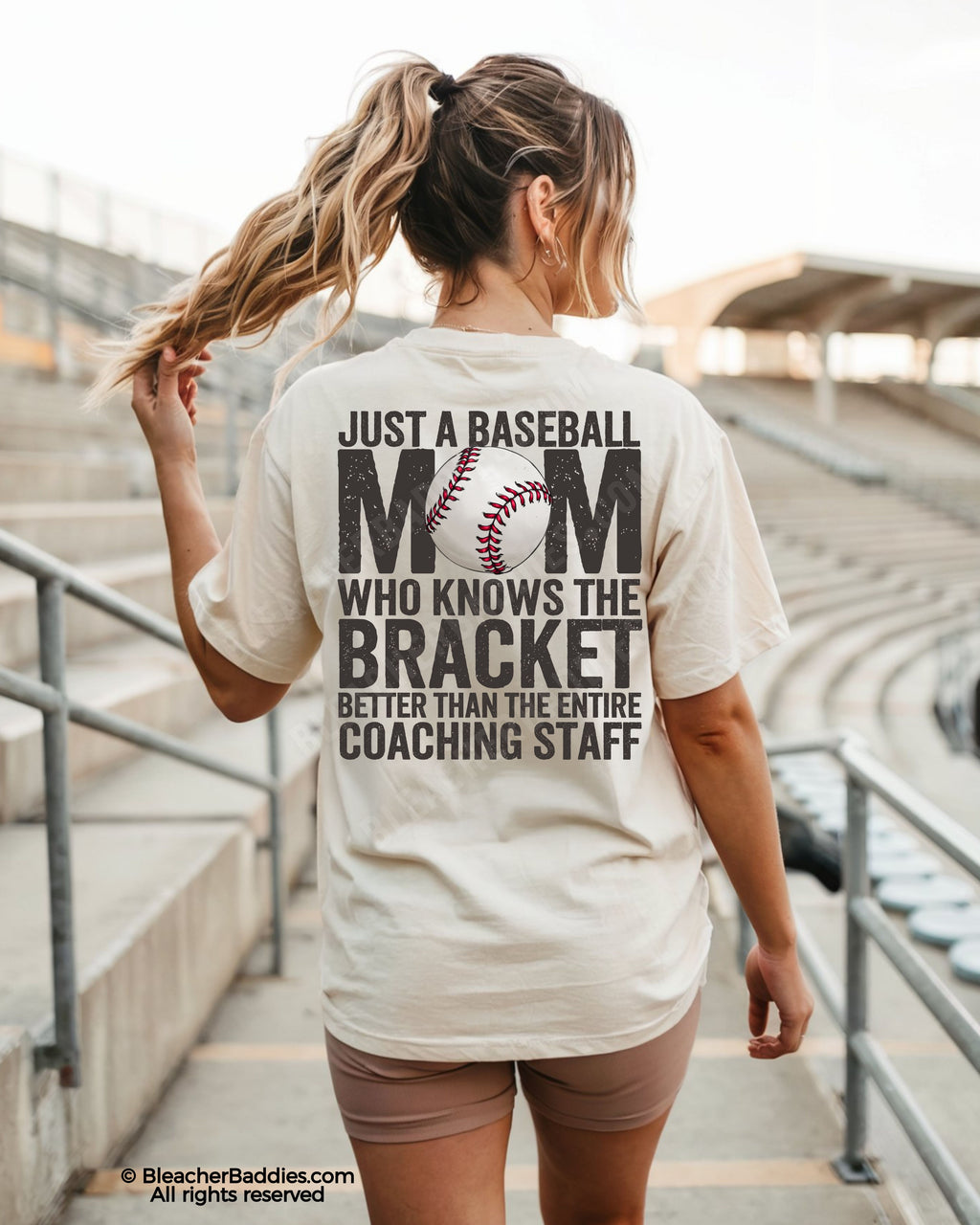 Woman wearing ivory baseball mom shirt in stadium with oversized distressed back graphic reading “Just a baseball mom who knows the bracket better than the entire coaching staff”