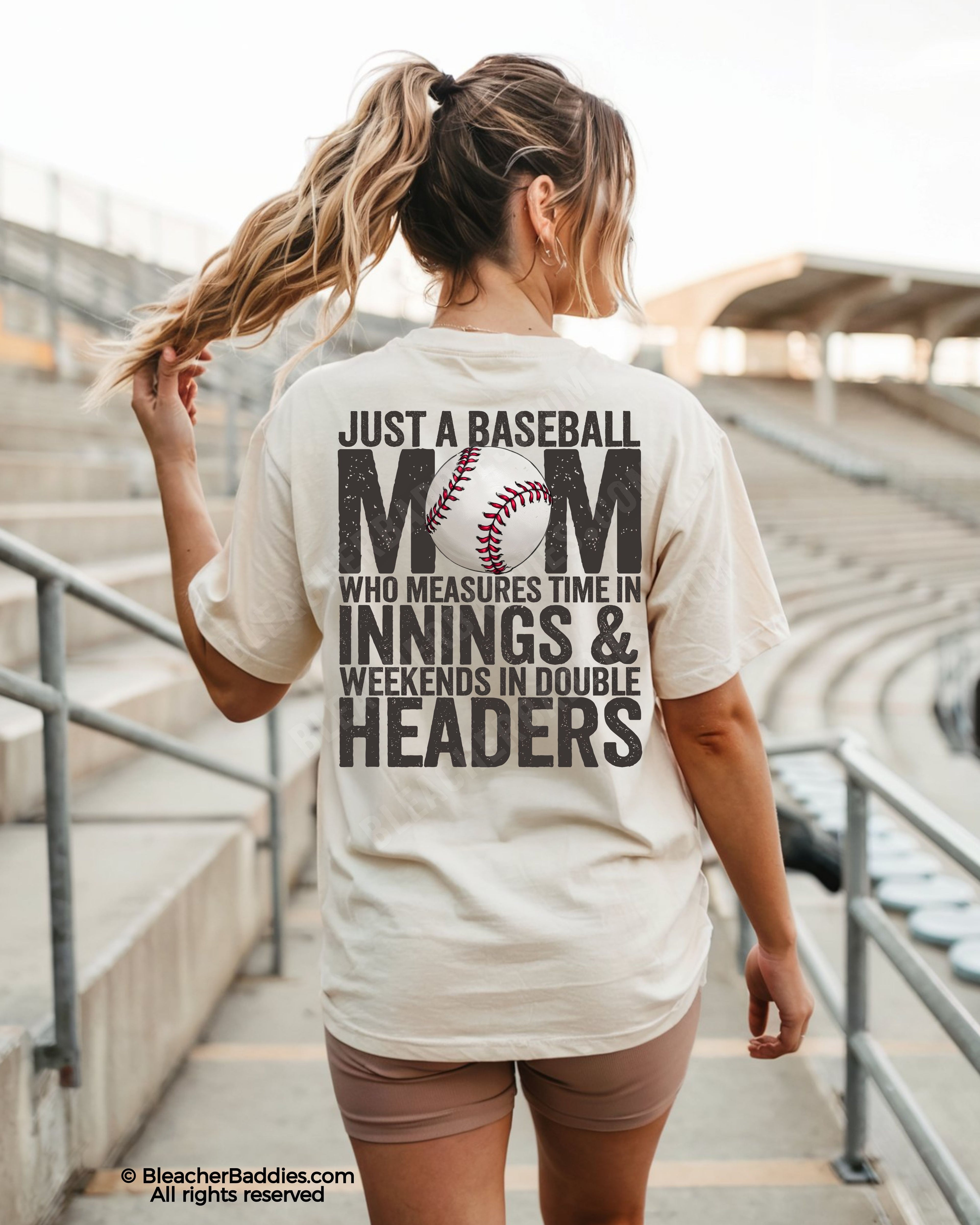 Woman walking through baseball stadium wearing ivory Comfort Colors game day shirt with back graphic reading “Just a Baseball Mom Who Measures Time in Innings & Weekends in Double Headers”
