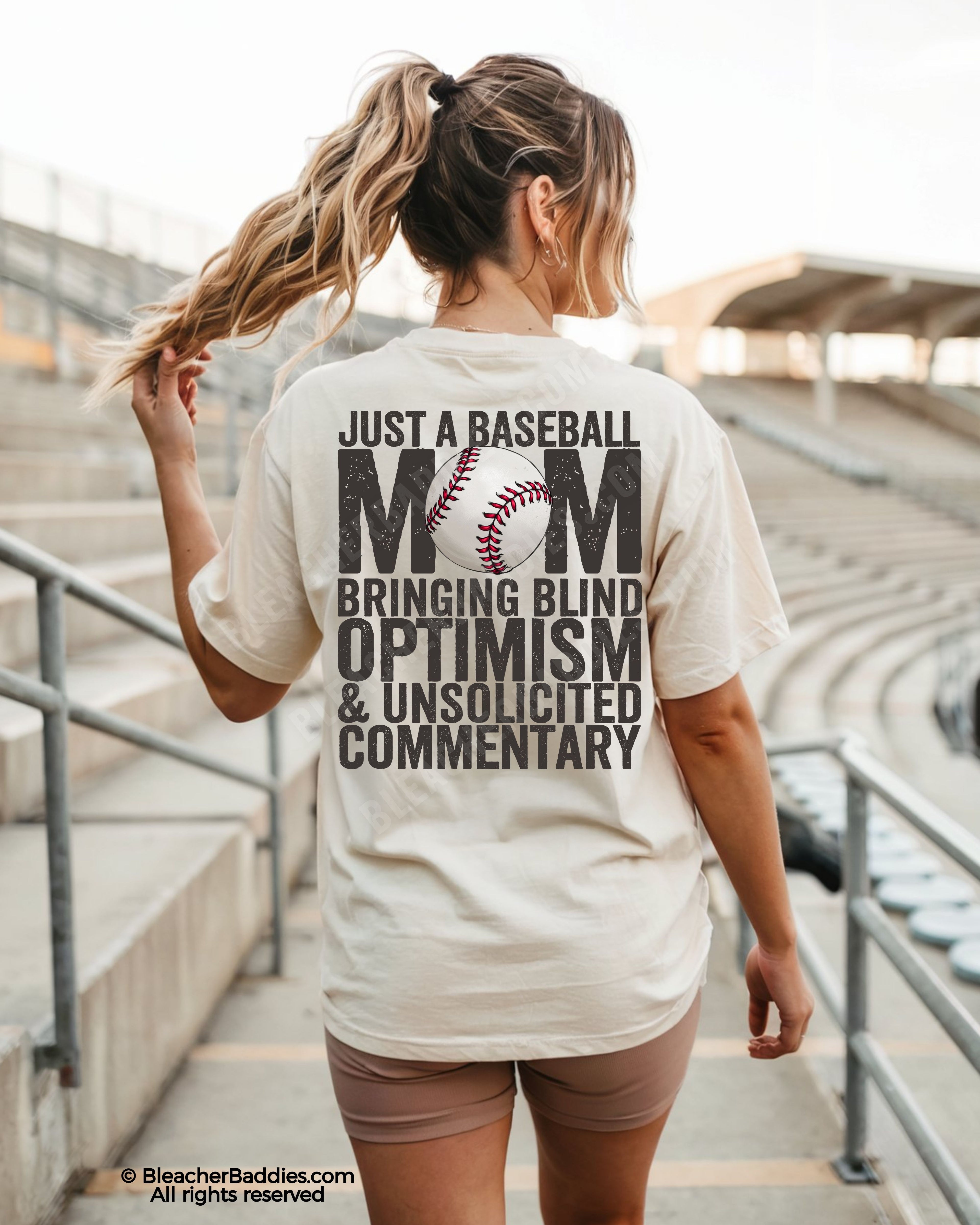 Woman walking through baseball stadium wearing ivory Comfort Colors game day shirt with back graphic reading “Just a Baseball Mom Bringing Blind Optimism & Unsolicited Commentary”