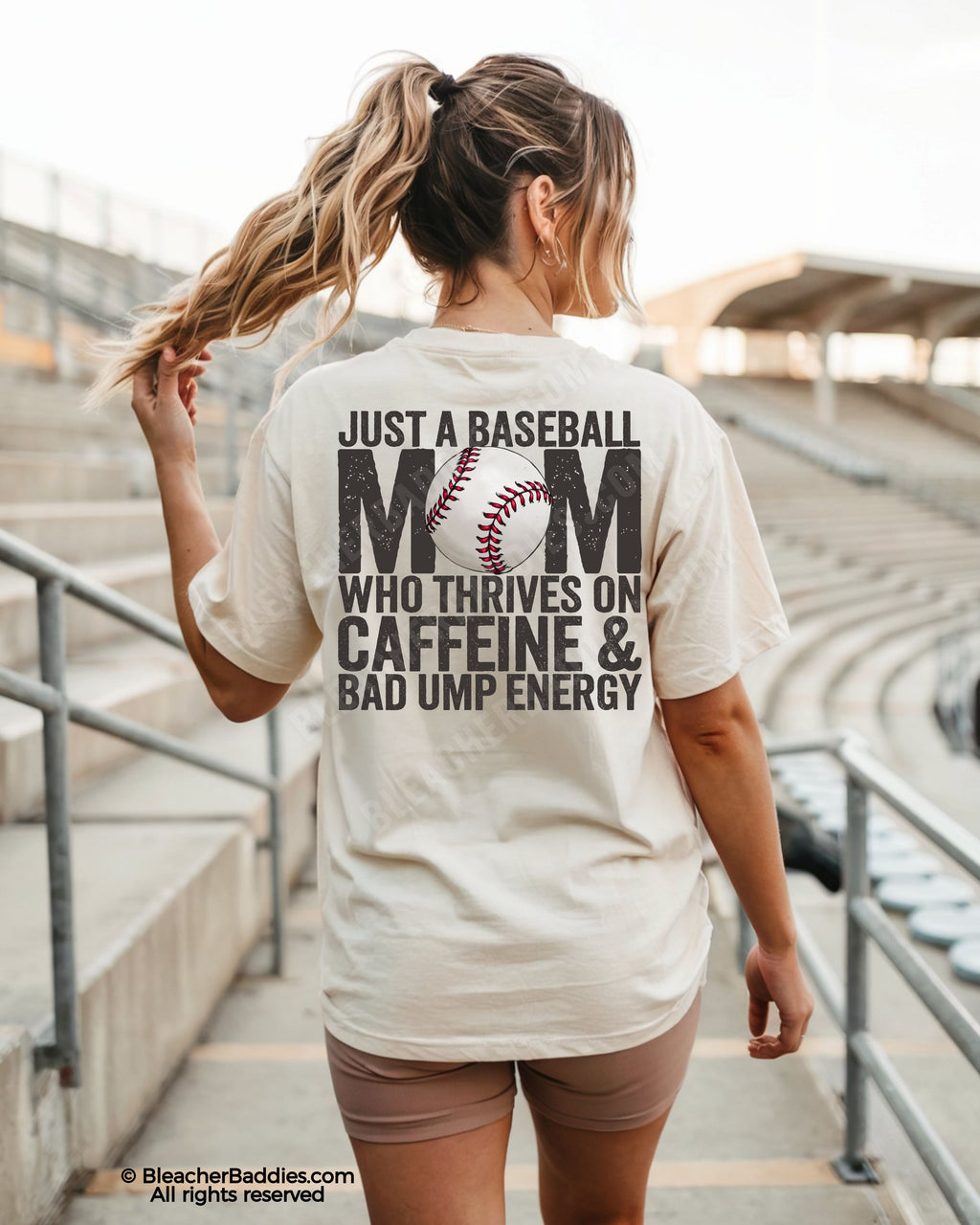 Woman walking through baseball stadium wearing ivory Comfort Colors baseball game day shirt with distressed back graphic reading “Just a Baseball Mom Who Thrives on Caffeine & Bad Ump Energy”