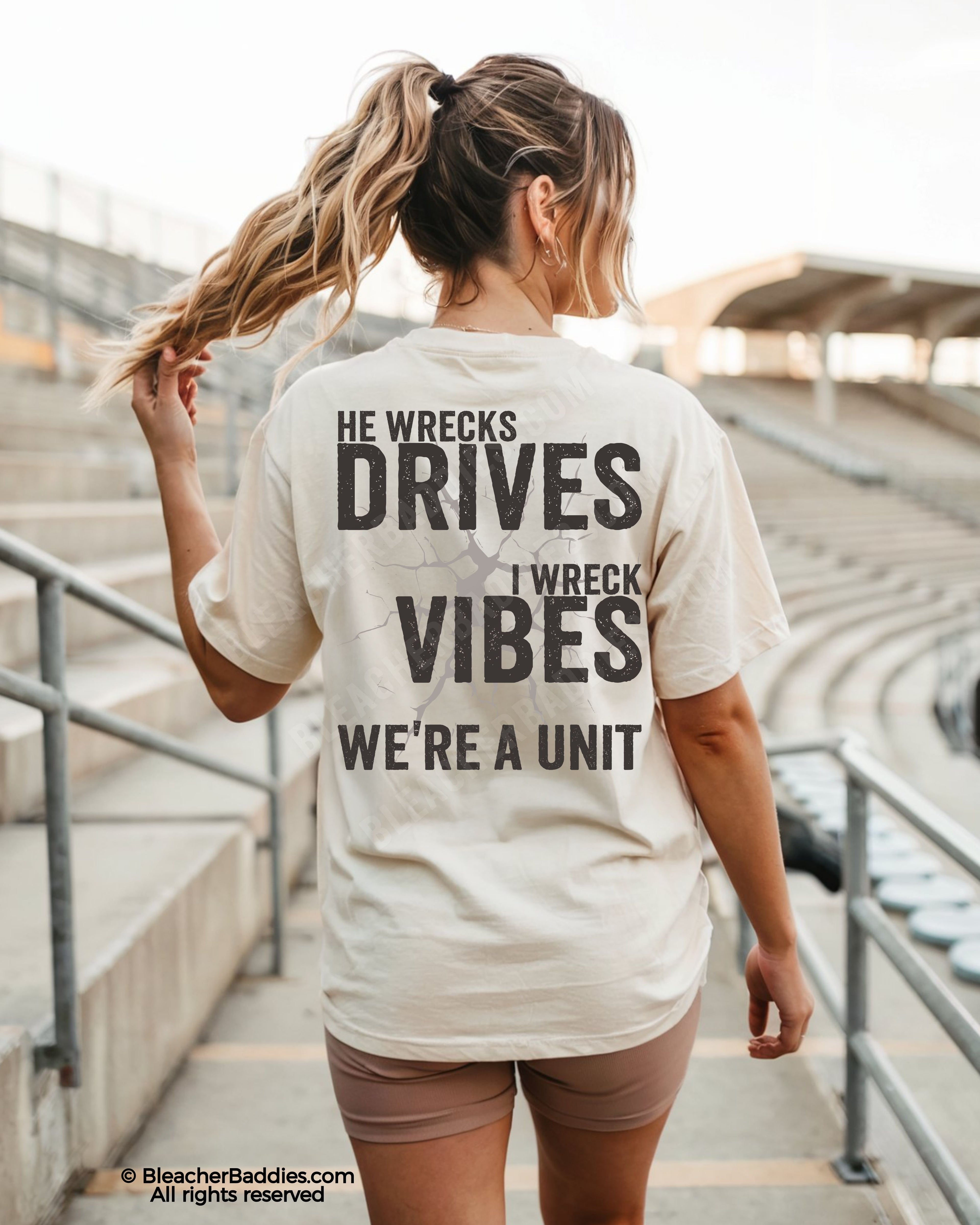 Woman walking through stadium stands wearing an ivory oversized t-shirt with a large distressed black back graphic that reads “He Wrecks Drives I Wreck Vibes We’re a Unit.”